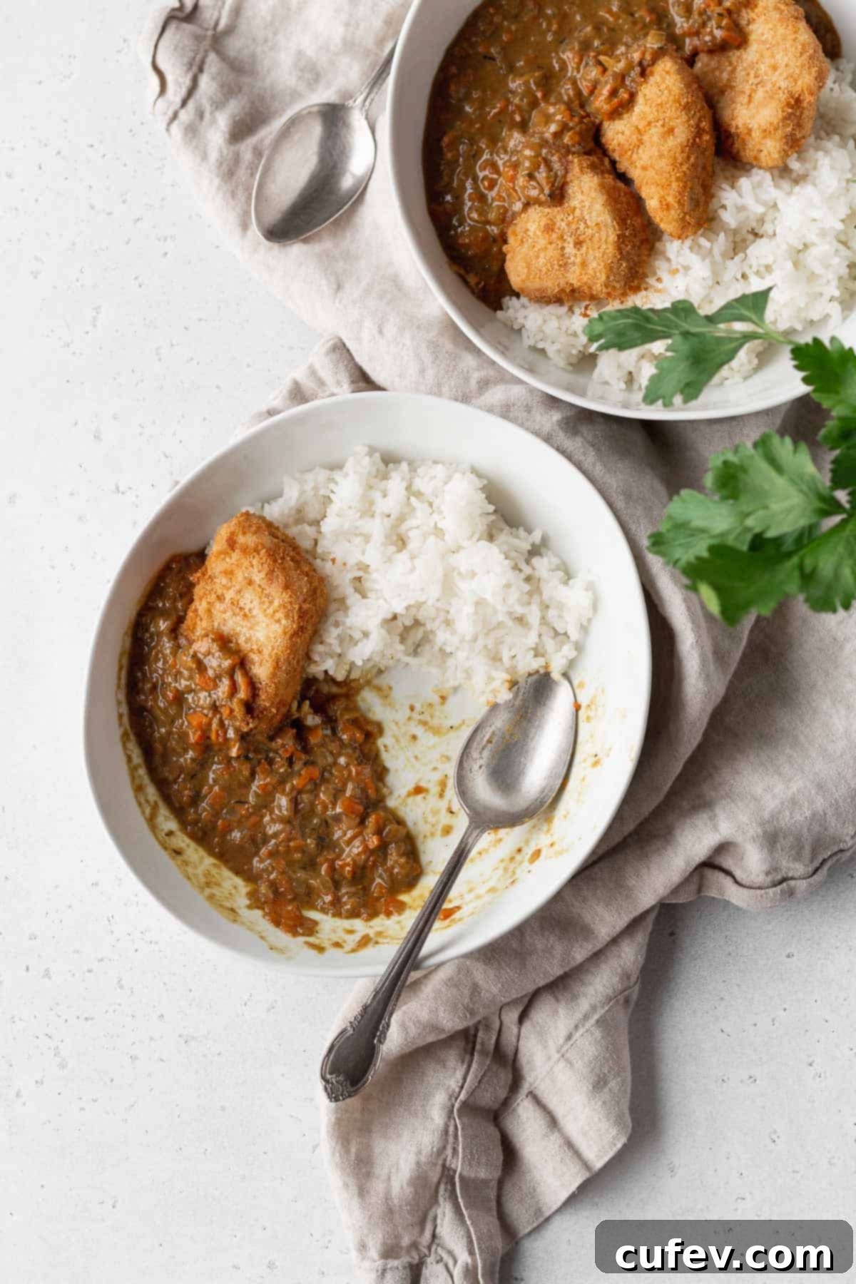 Two enticing bowls of vegan katsu curry, elegantly presented on a natural linen fabric alongside a gleaming silver spoon, inviting you to dig in.