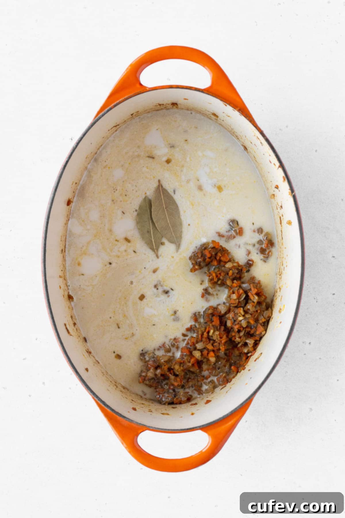 Creamy coconut milk and savory vegetable stock being poured over the aromatic vegetables, with two bay leaves resting on top, ready for simmering.