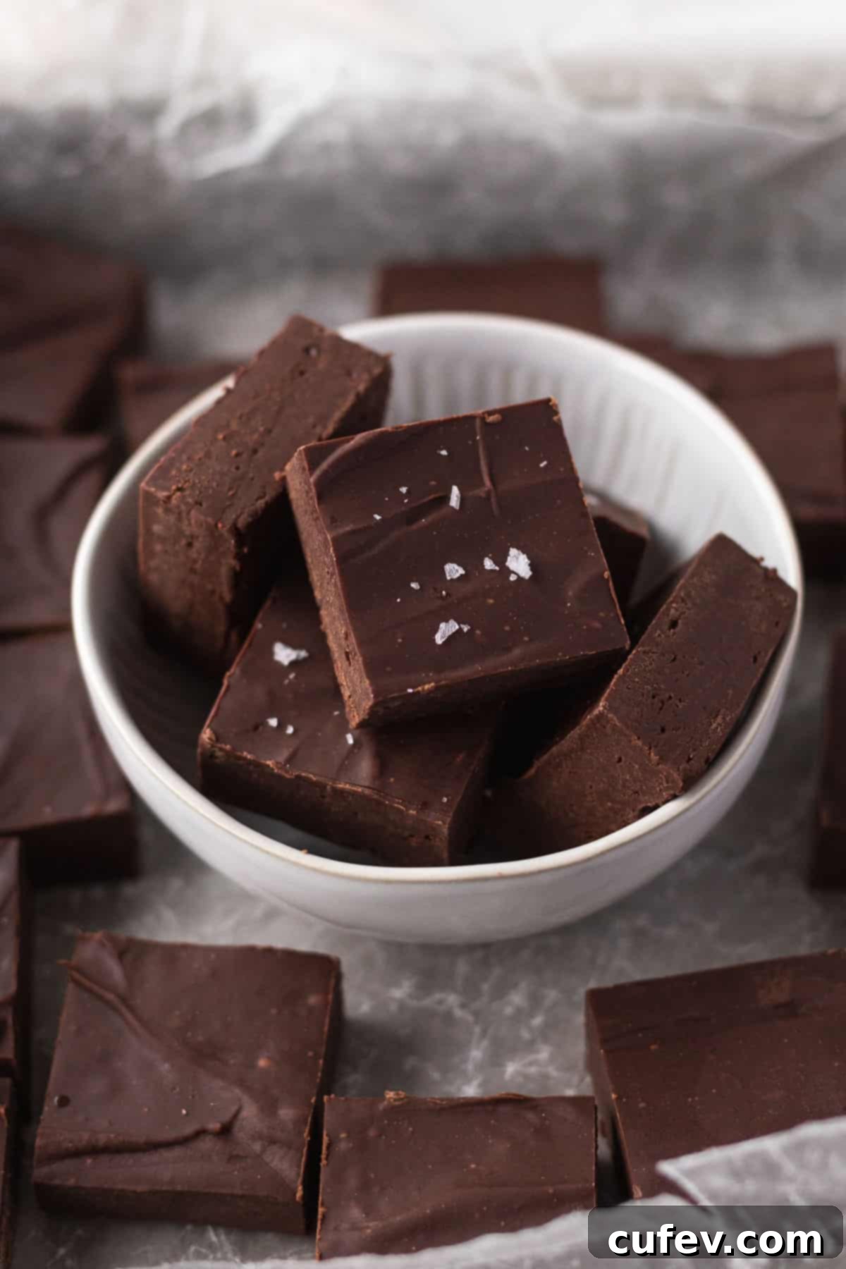 45 degree angle shot of cubes of dairy-free fudge in a white bowl on a piece of crumpled white parchment with other cubes of fudge.