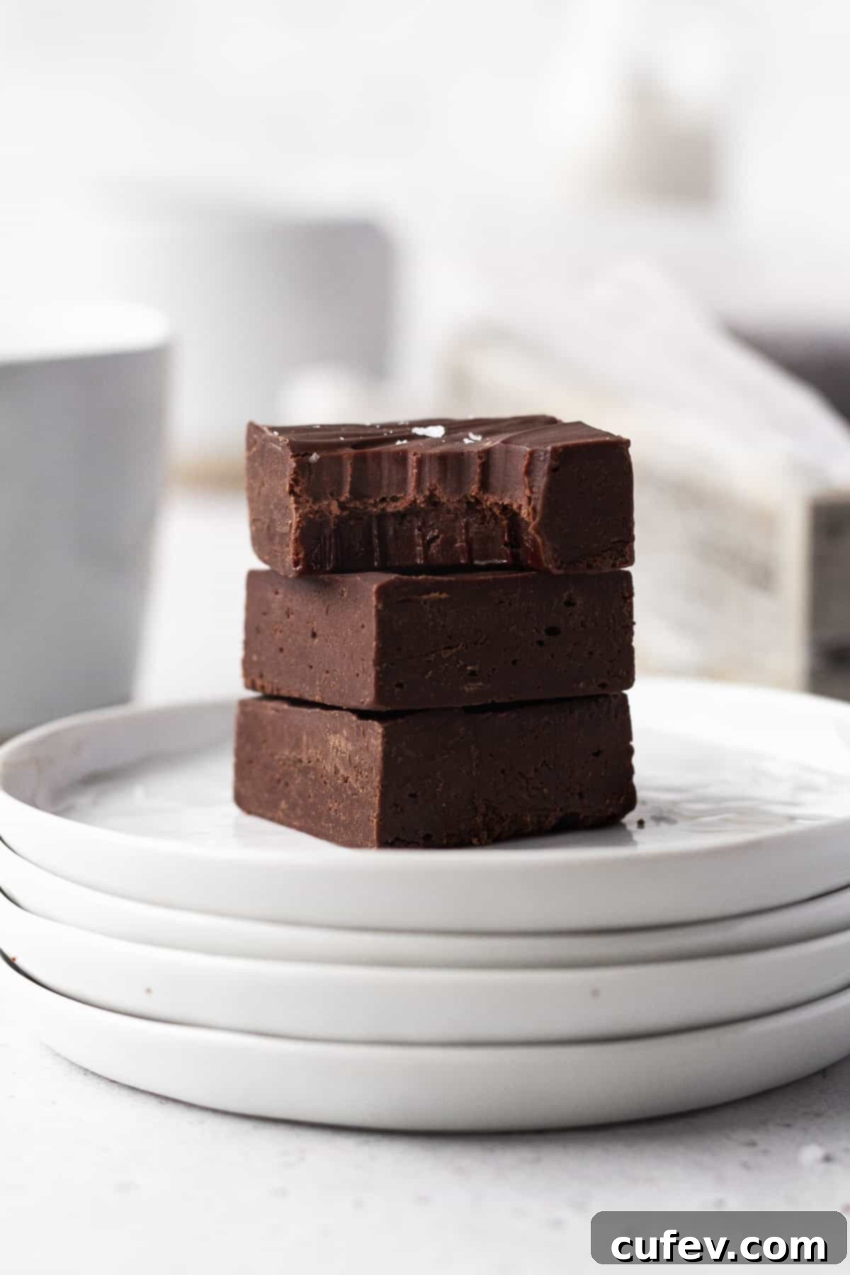 Side on shot of 3 cubes of dairy-free fudge stacked on a stack of white plates with the top square having a bite taken out.