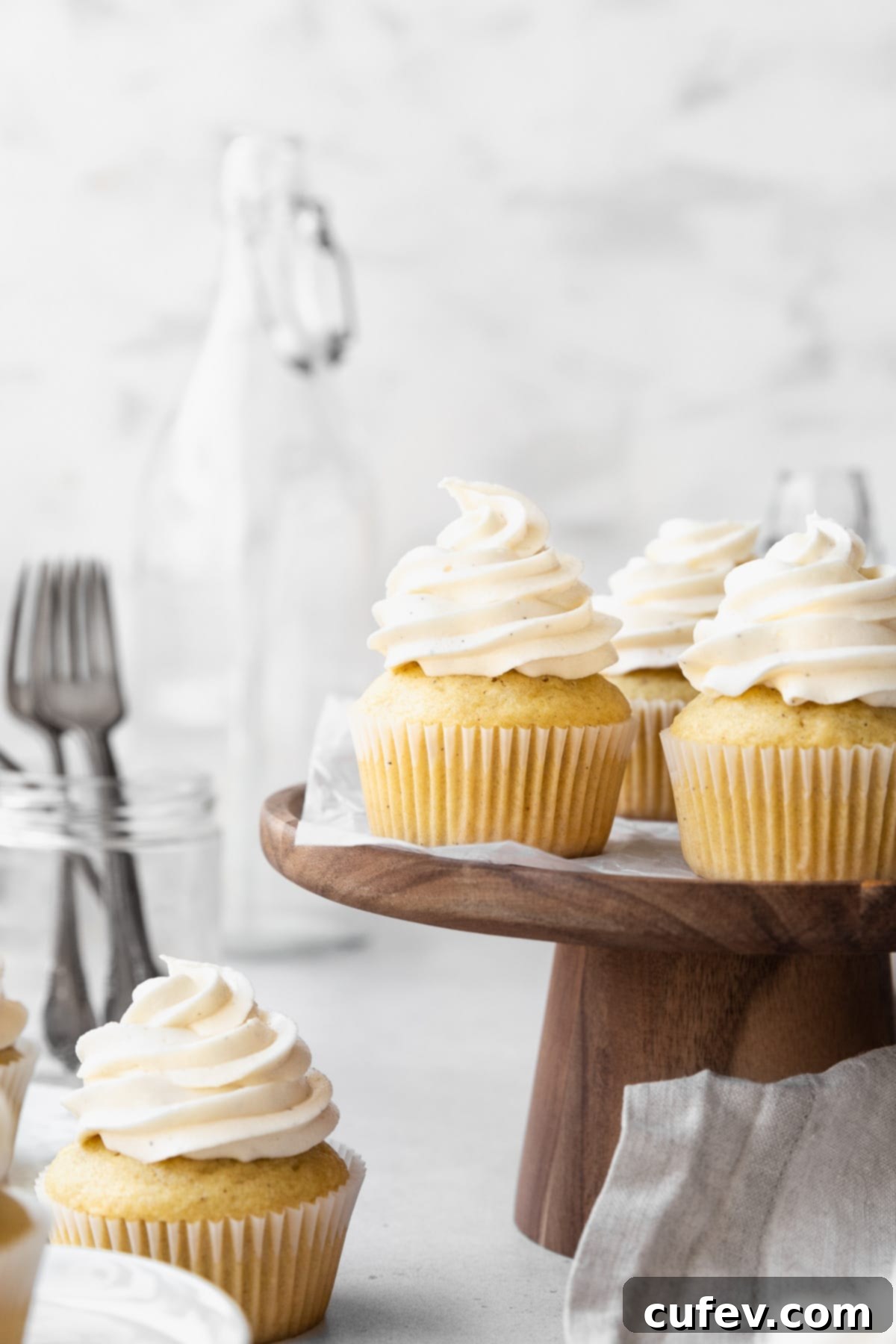 Guilt-Free Cupcakes 2 Side on shot of dairy-free vanilla cupcakes on a wooden cake stand with a jar of forks and a large jug in the background. The cupcakes are perfectly baked and topped with smooth, white frosting.