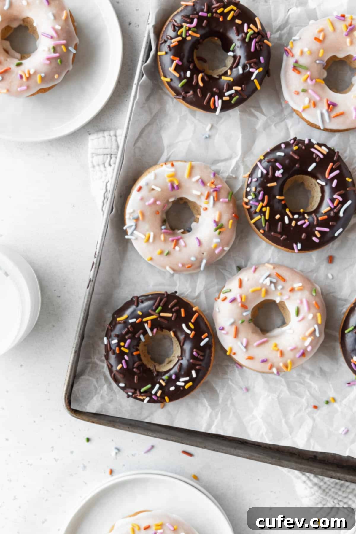 Overhead shot of a parchment lined tray of chocolate and vanilla dipped vegan baked donuts on a white table with small dessert plates holding vanilla glazed vegan doughnuts.