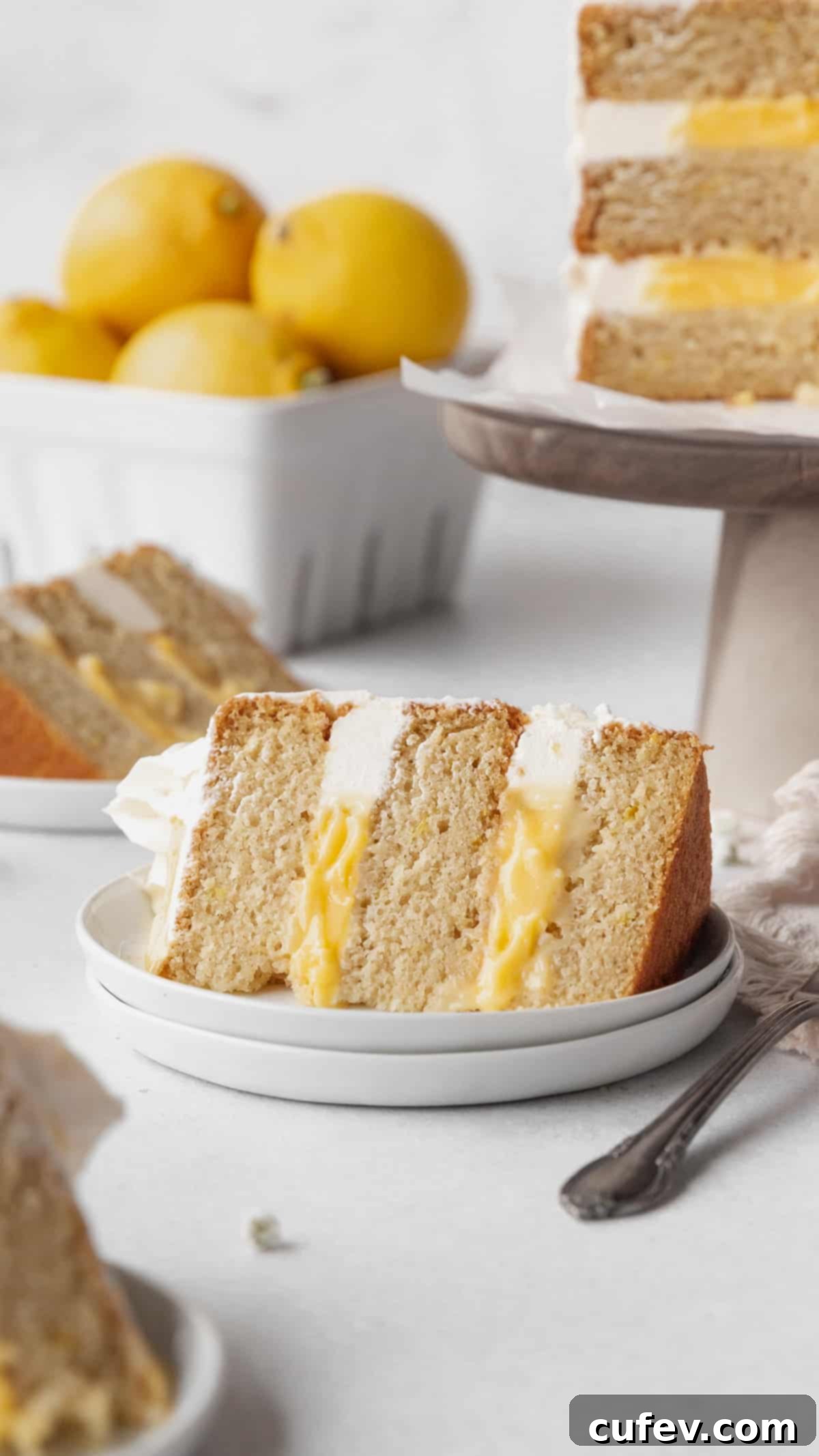 A slice of lemon curd cake on a white dessert plate with a cake stand holding the rest of the cake and a ceramic fruit pint holding a handful of lemons in the background.