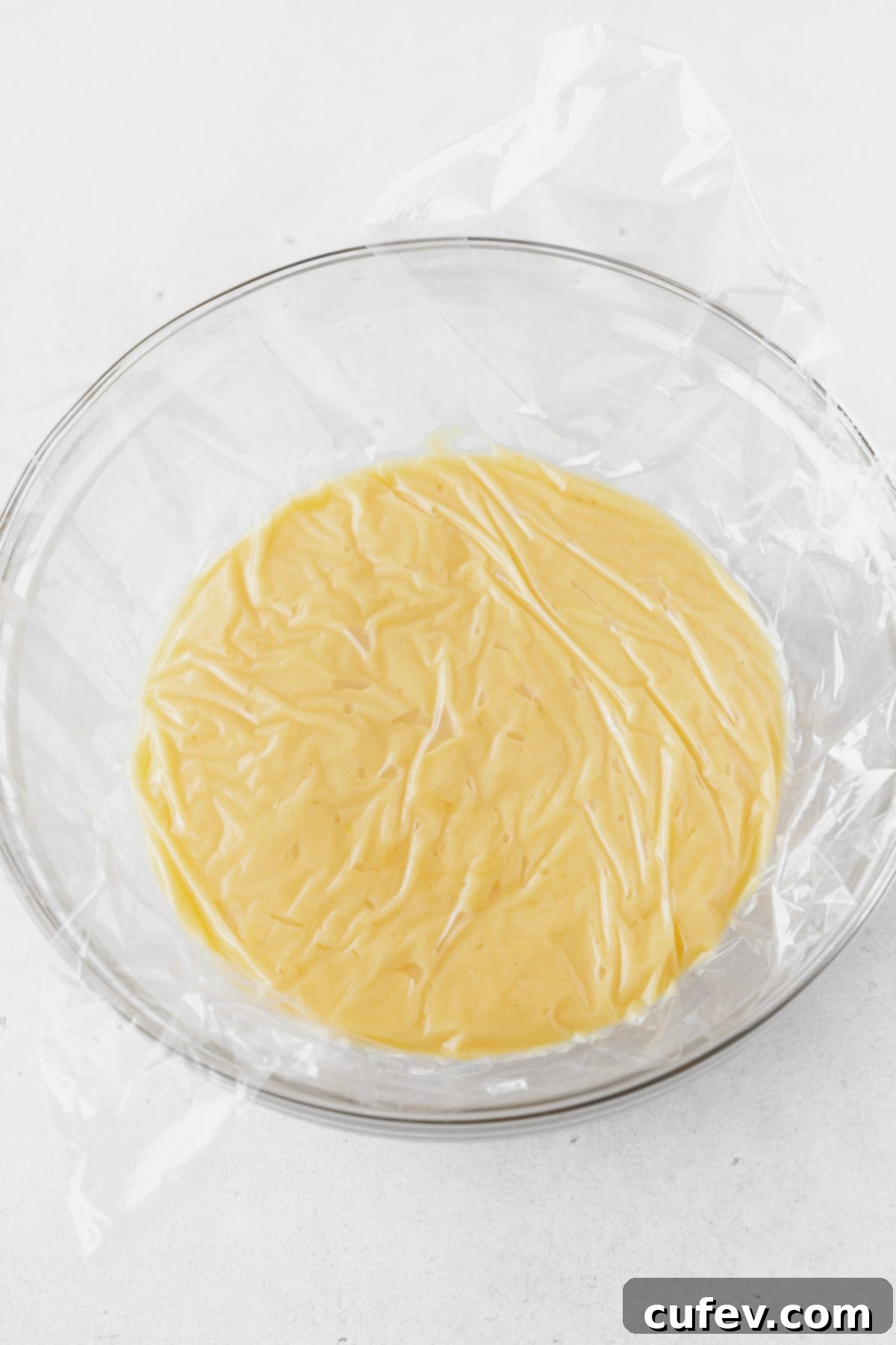 A glass bowl of lemon curd with a plastic wrap cover against the surface.