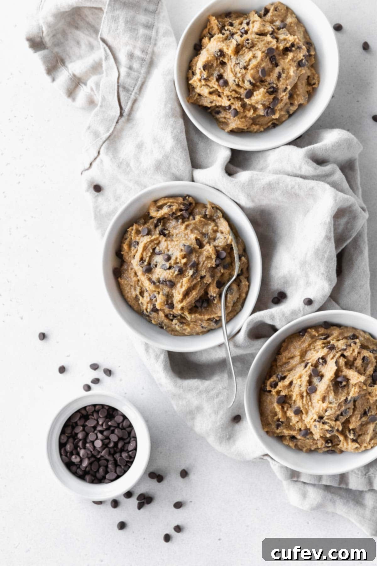 Overhead hero shot of 3 bowls of gluten-free cookie dough with a silver spoon in the middle bowl, and a small bowl of mini chocolate chips to the side.