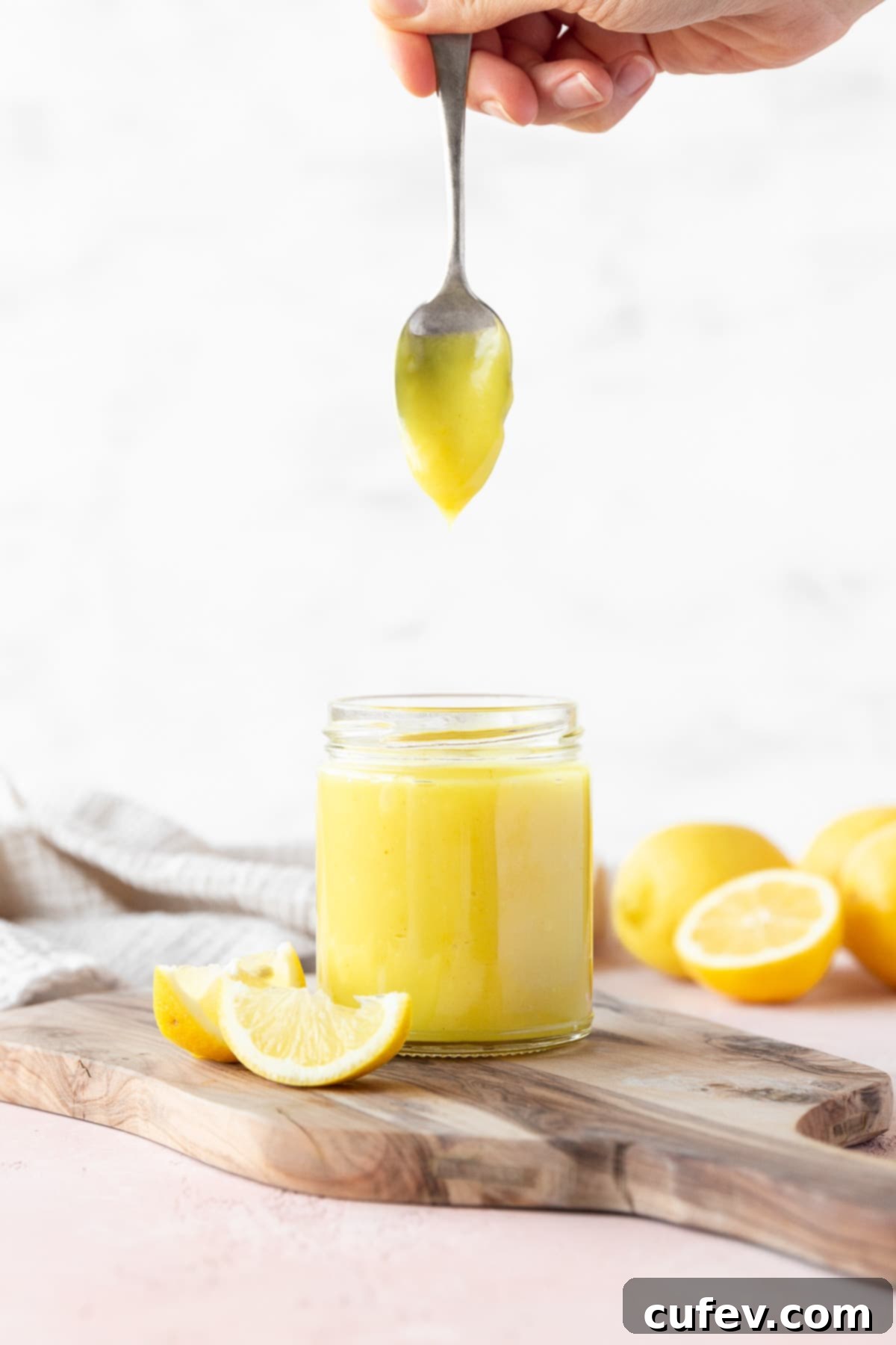 Velvety Vegan Lemon Curd 2 Side on shot of a hand holding a spoon of vegan lemon curd vertically over the full mason jar with the curd slowly dripping off, showcasing its creamy consistency.