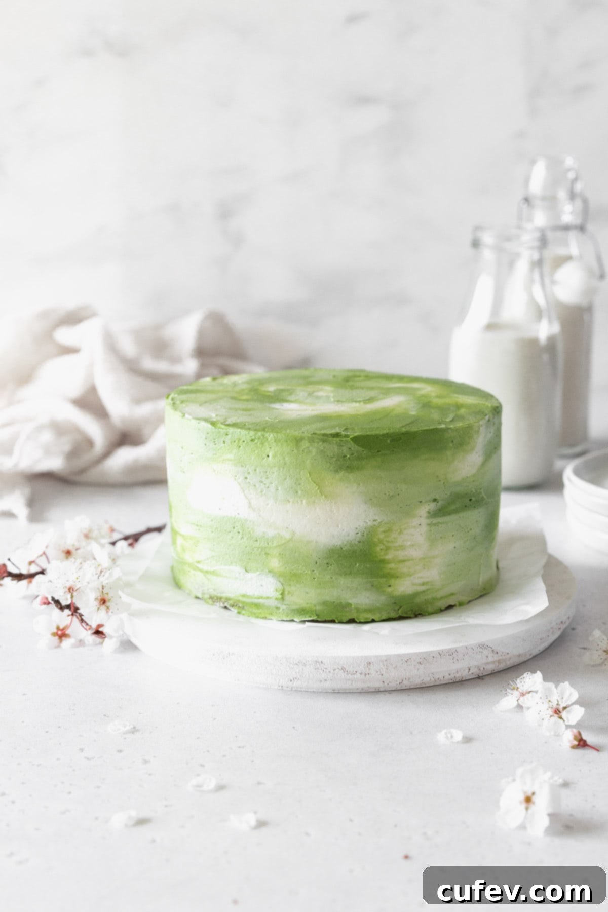 An iced matcha cake on a white cake stand with cherry blossoms. 