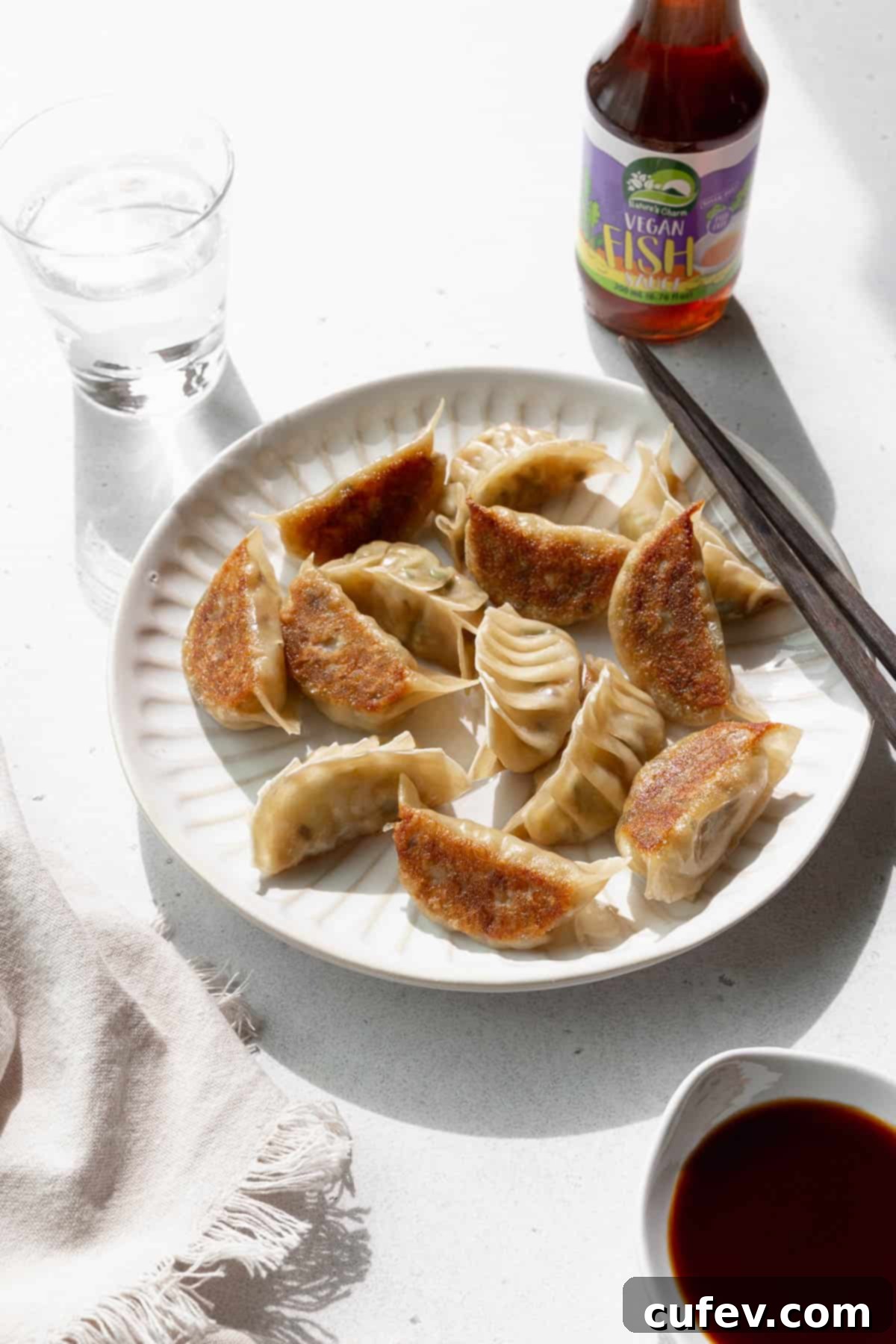 Angled shot of vegetable gyoza on a white plate with a glass of water and a bottle of vegan fish sauce in the background.