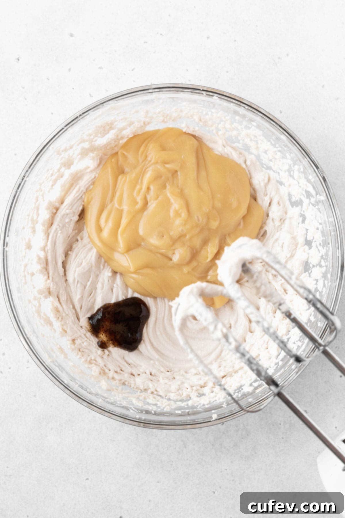Sweetened condensed oat milk and vanilla extract being added to the whipped oat cream in a glass mixing bowl, blending into a creamy ice cream base.