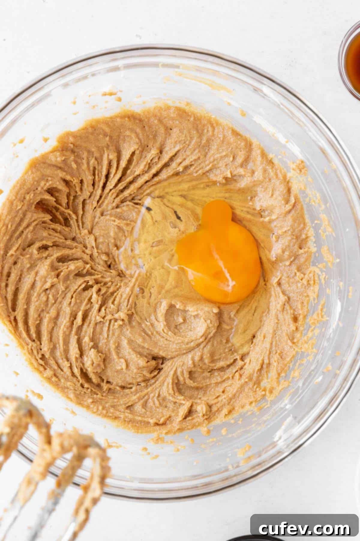 Adding an egg to the creamed peanut butter and vegan butter mixture in the mixing bowl, followed by vanilla extract for binding and flavor.