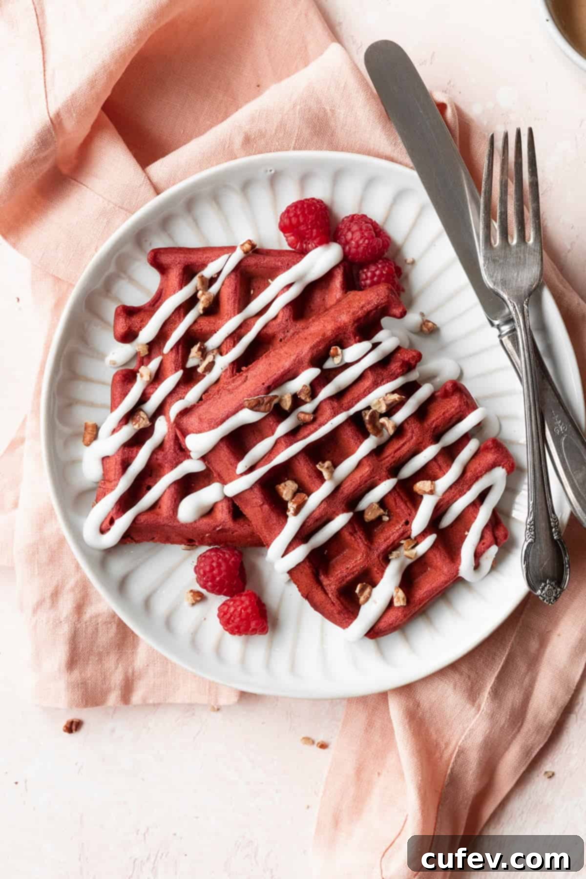 A closeup shot of a white plate of red velvet waffles with cream cheese glaze and raspberries.