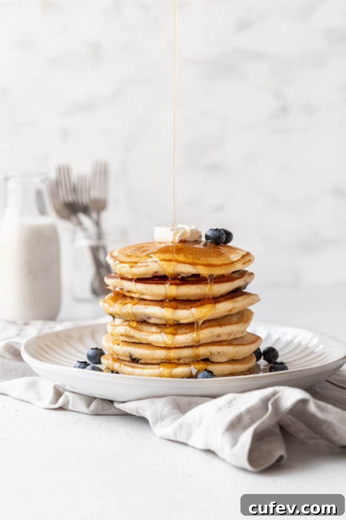A short stack of blueberry studded muffin mix pancakes being drizzled with syrup, capturing a dynamic side-on action shot.
