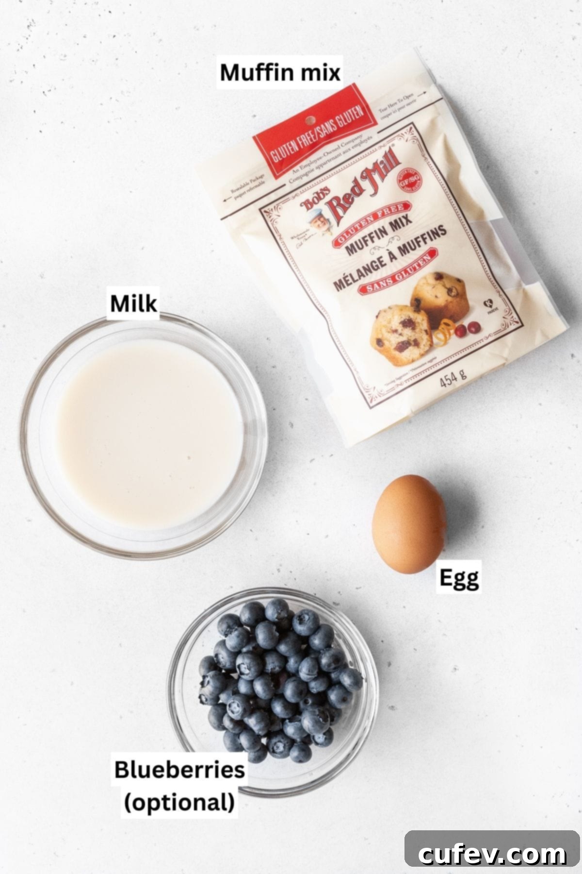 Bowls of muffin mix, a cracked egg, and milk, along with a small dish of blueberries, are neatly arranged on a white table, showing the simple ingredients needed for muffin mix pancakes.