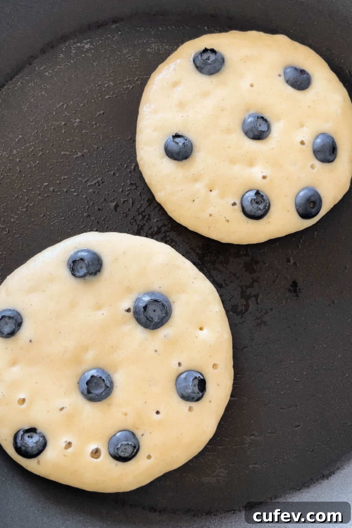 A process shot showing 6-8 fresh blueberries being added to each pancake on the griddle just after pouring the batter.