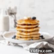 An action shot of syrup being poured onto a delectable stack of blueberry muffin mix pancakes, crowned with a pat of butter and garnished with extra fresh blueberries.