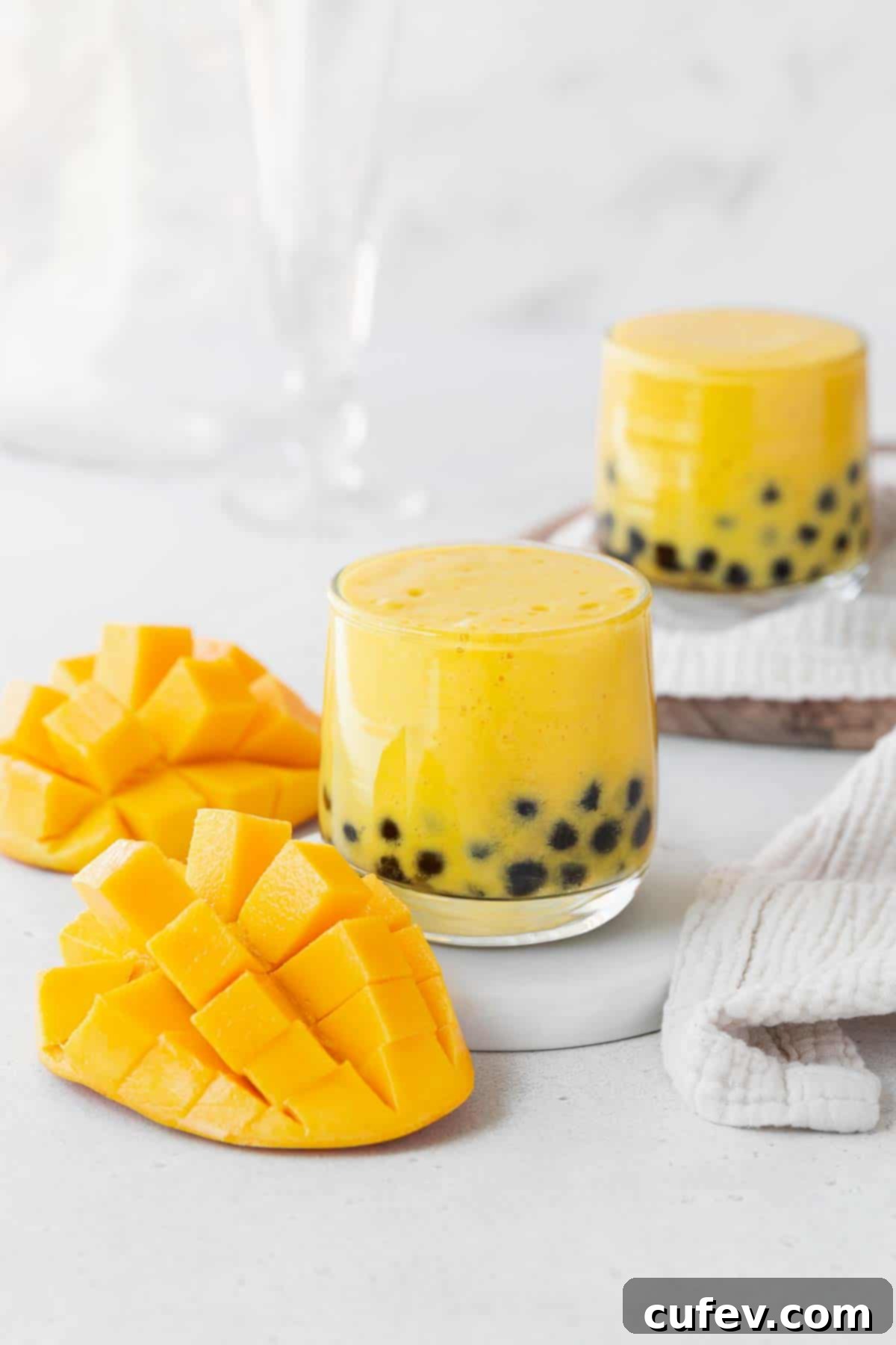 Tropical Mango Bubble Tea 2 Two glasses of mango boba tea on a white counter next to a cubed up mango, showcasing the vibrant colors and appealing texture.