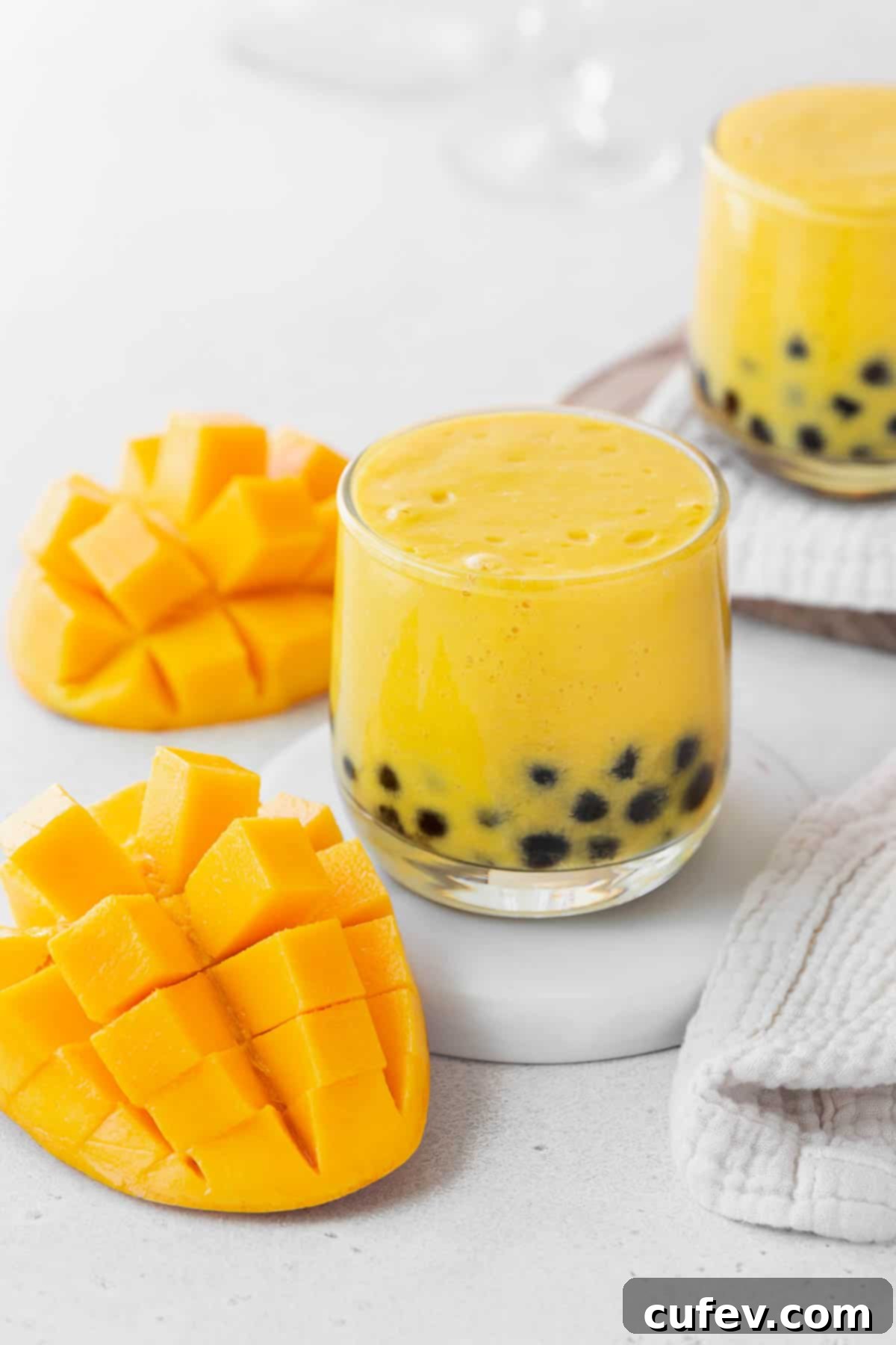 Tropical Mango Bubble Tea 4 A closeup shot of a small glass of mango boba tea on a white coaster on the counter next to a sliced up mango, emphasizing the drink's texture and color.