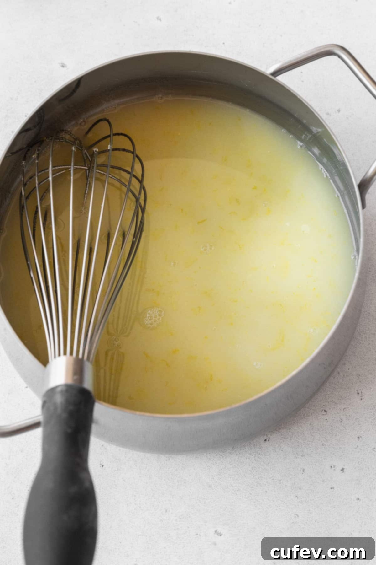 A saucepan filled with a slightly thickened mixture of whisked lemon juice, zest, and sugar, gently heating on the stovetop.