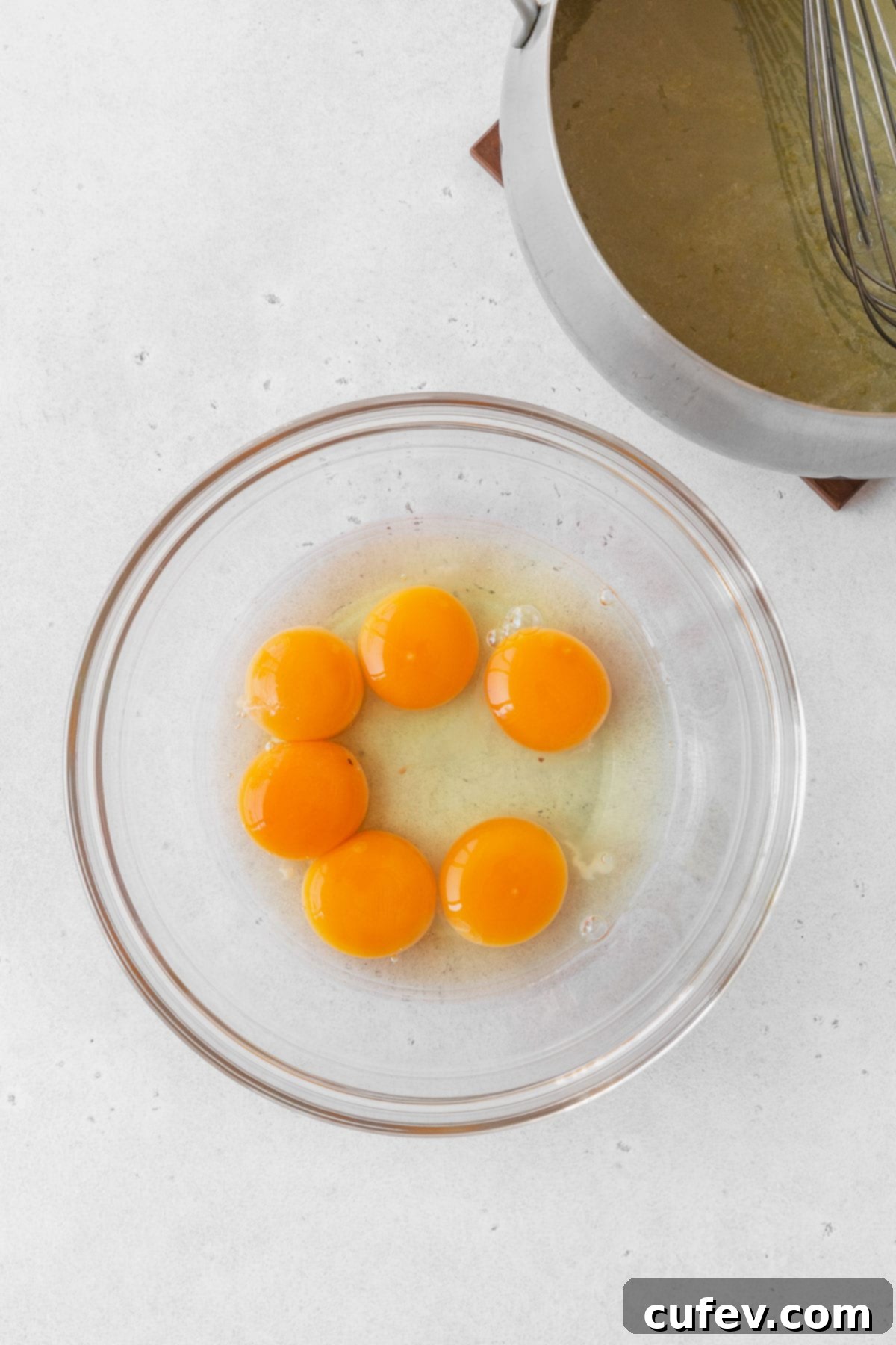 Three whole eggs and three separate egg yolks in a glass mixing bowl, poised for whisking into a smooth consistency.