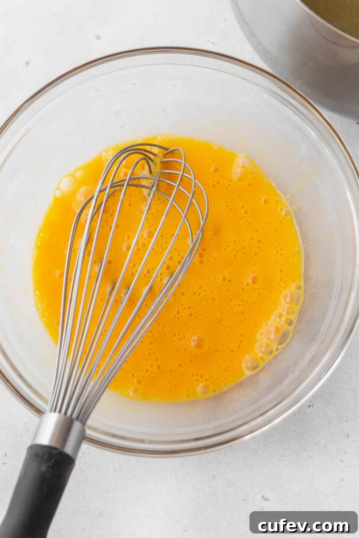 Beautifully whisked egg yolks and whole eggs in a transparent glass bowl, with a whisk resting inside, showing a smooth, unified mixture.