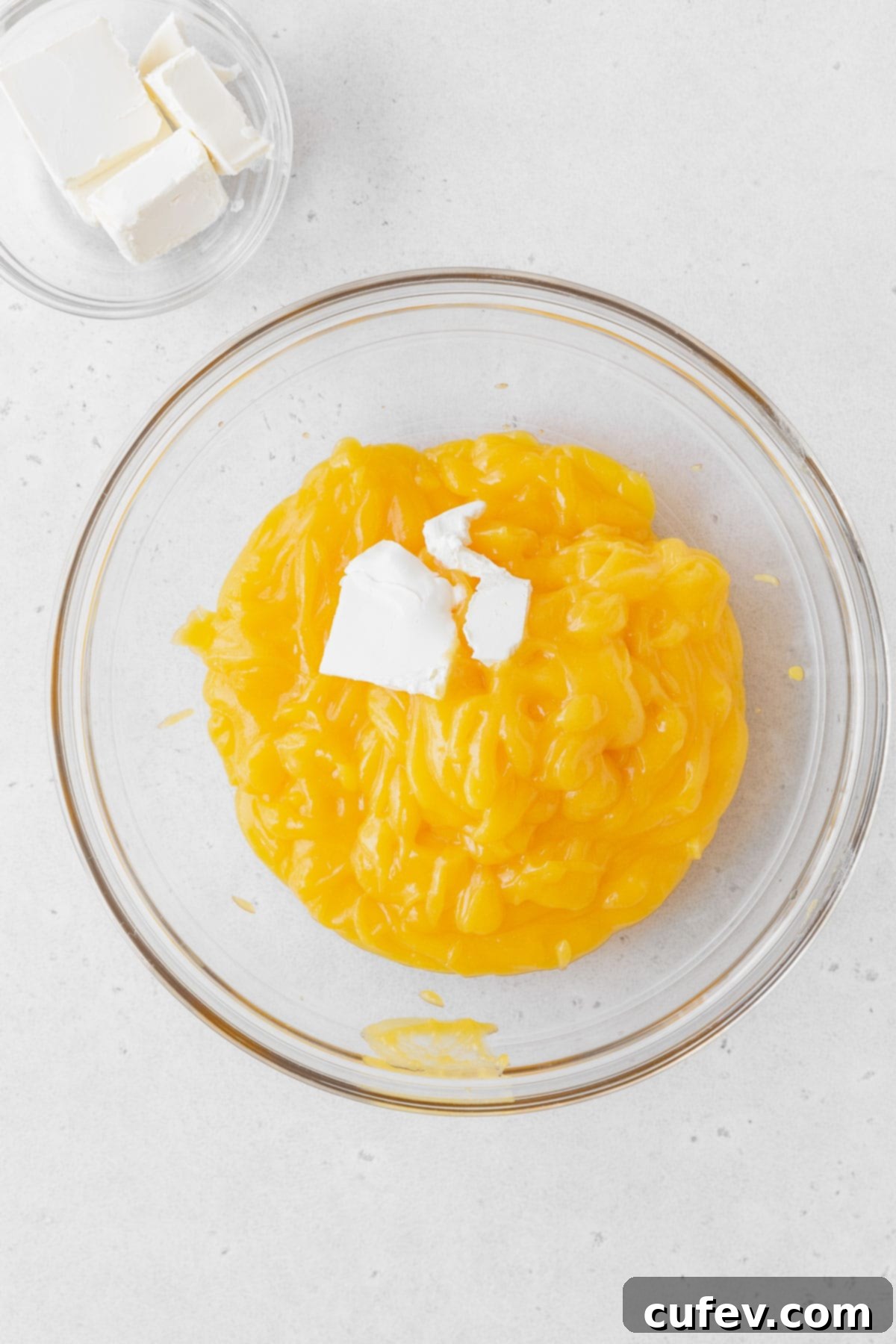 Adding individual cubes of vegan butter to the warm, freshly sieved lemon curd in a mixing bowl.