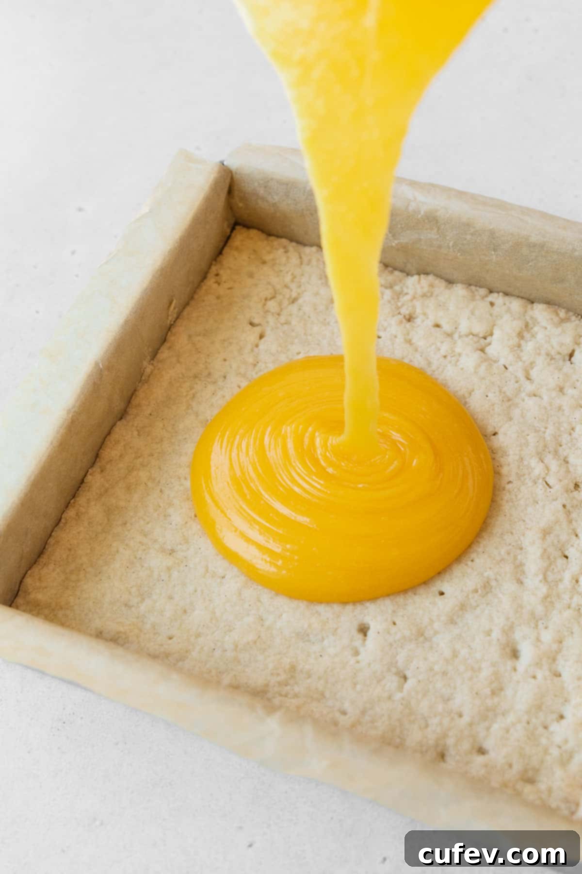The glistening dairy-free lemon curd filling being poured over the golden-brown shortbread crust in a baking pan.