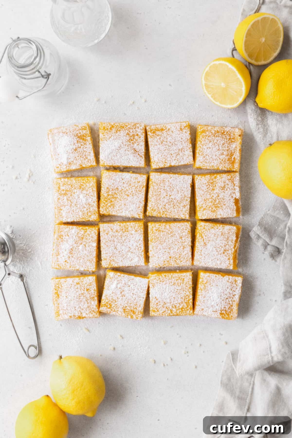A pan of perfectly cut dairy-free lemon bars arranged on a counter, accompanied by halved lemons, a dusting of powdered sugar, and a clean napkin, ready to be served.