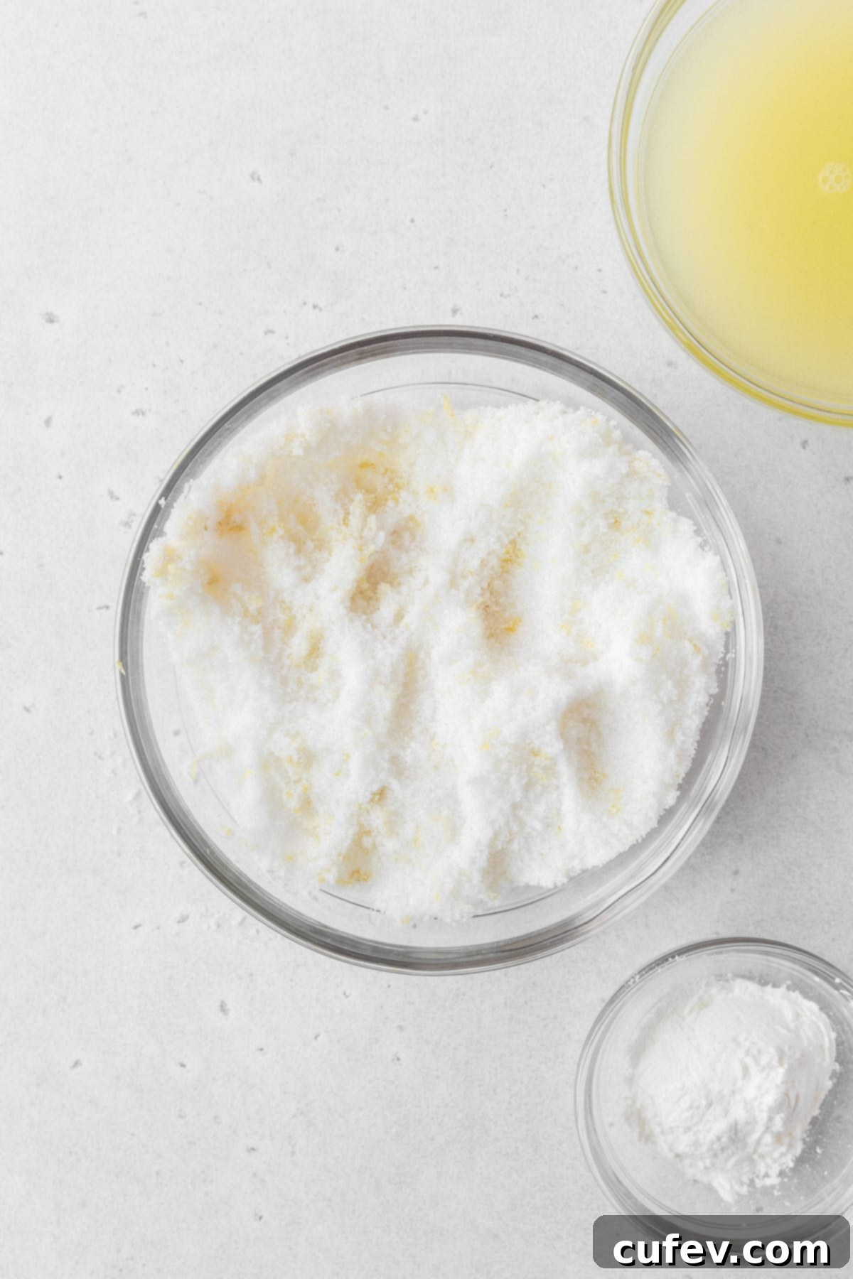 A clear glass mixing bowl containing granulated sugar intimately mixed with vibrant yellow lemon zest.