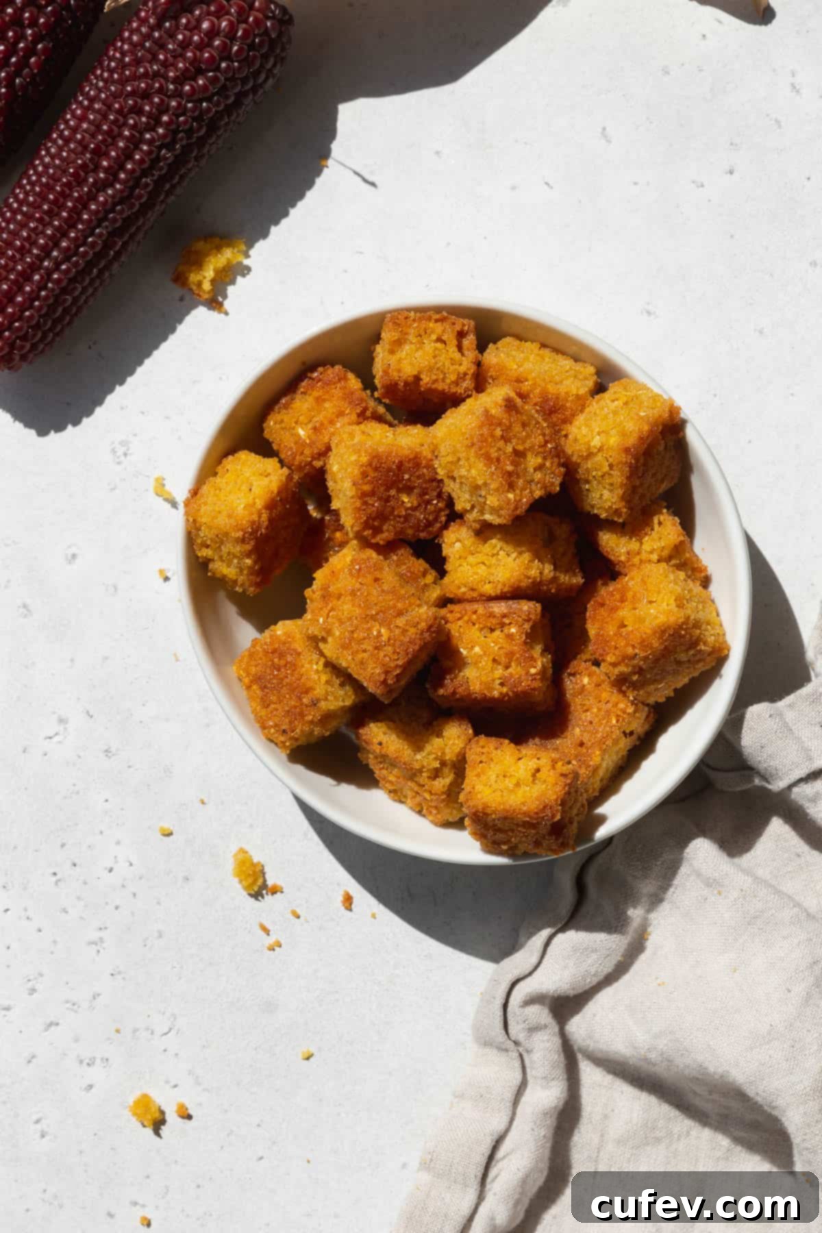 An overhead shot of a small bowl of golden-brown cornbread croutons with a napkin and a stalk of heirloom corn, emphasizing their crisp texture.