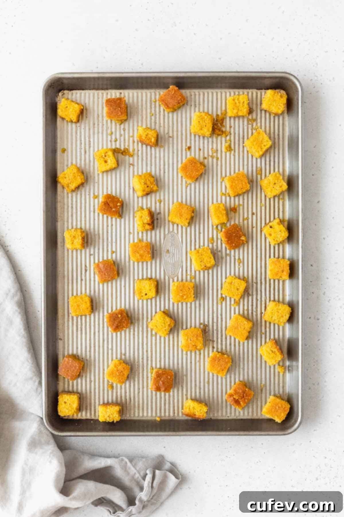 The seasoned cornbread croutons spread out in a single layer on a rimmed cookie sheet, poised for baking to achieve maximum crispness.