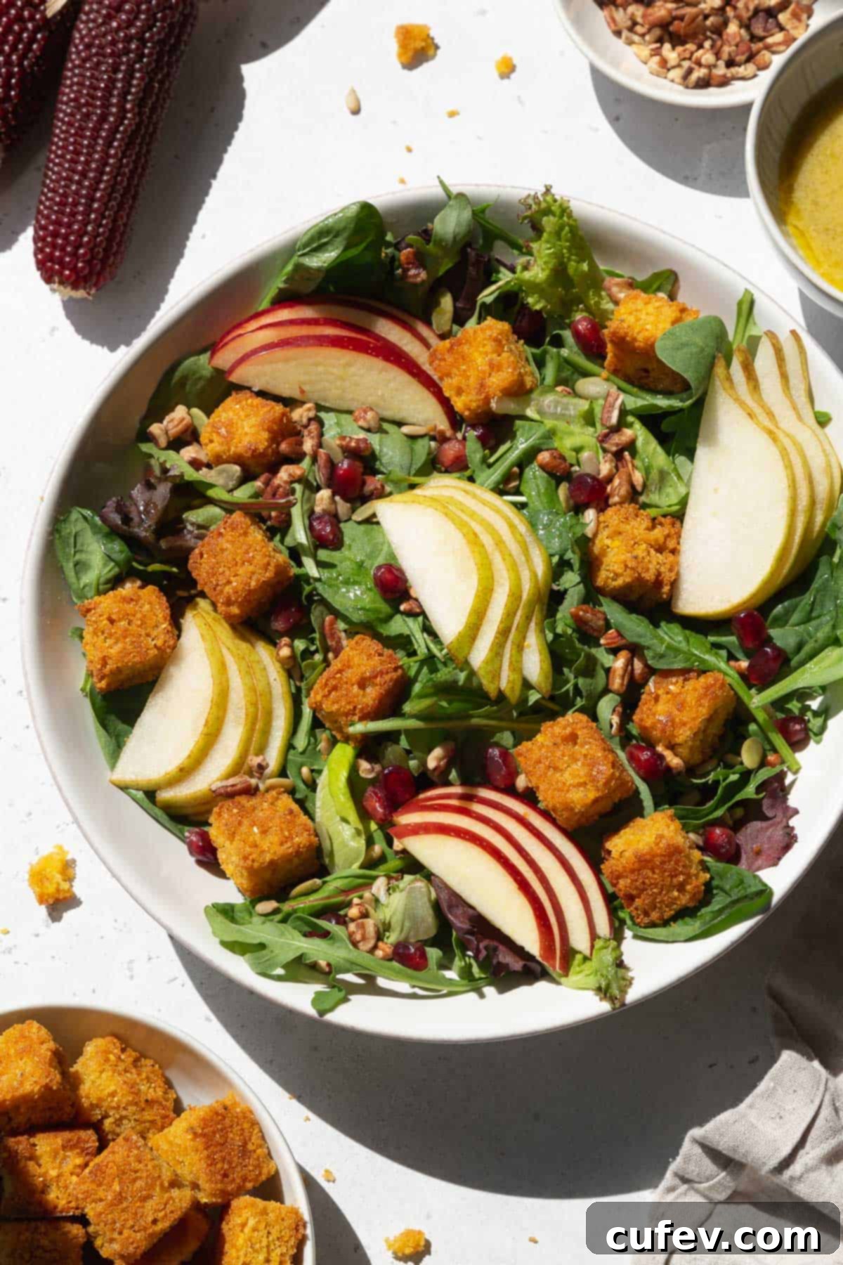 An overhead shot of a large white bowl of fresh salad, beautifully topped with golden cornbread croutons, vibrant sliced pears and apples, and glistening pomegranate seeds.