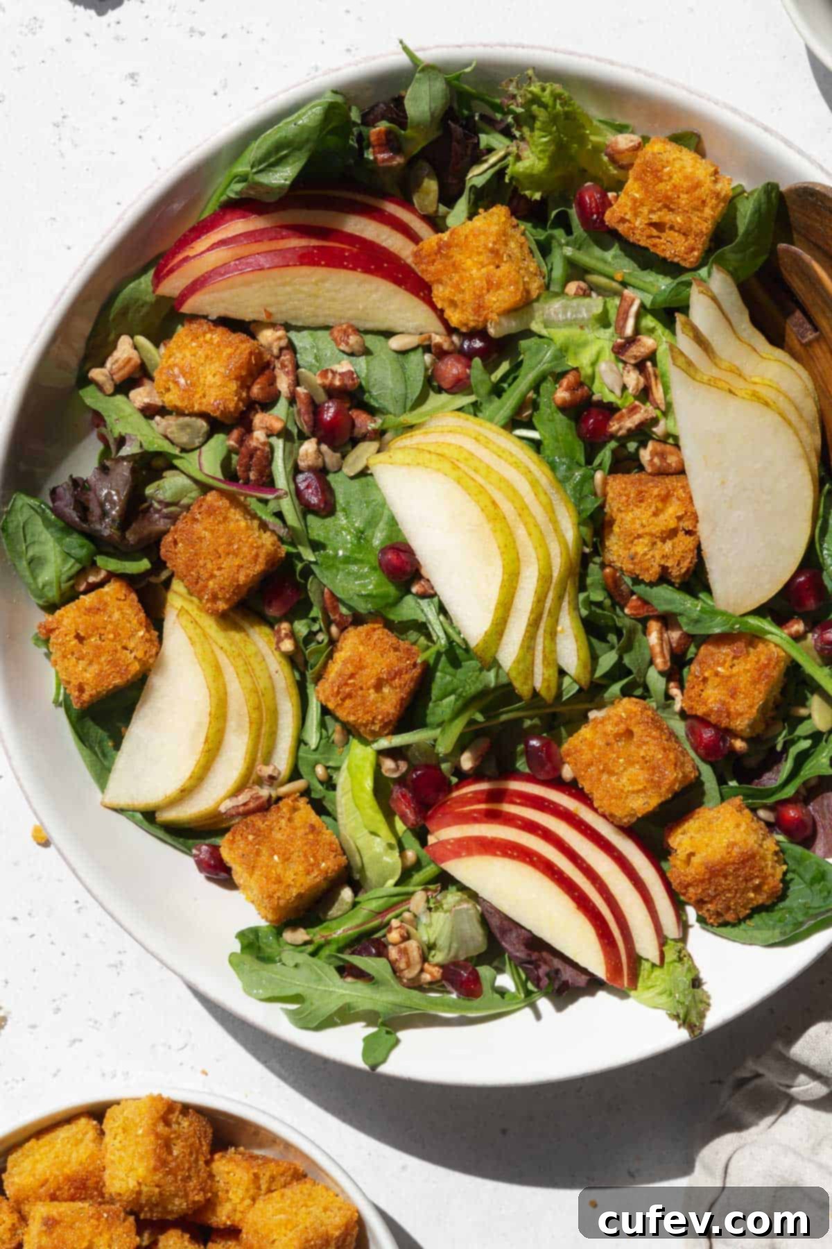 A large, inviting bowl of fresh salad, brimming with vibrant greens, crisp sliced apples, succulent pears, and an ample topping of golden cornbread croutons.