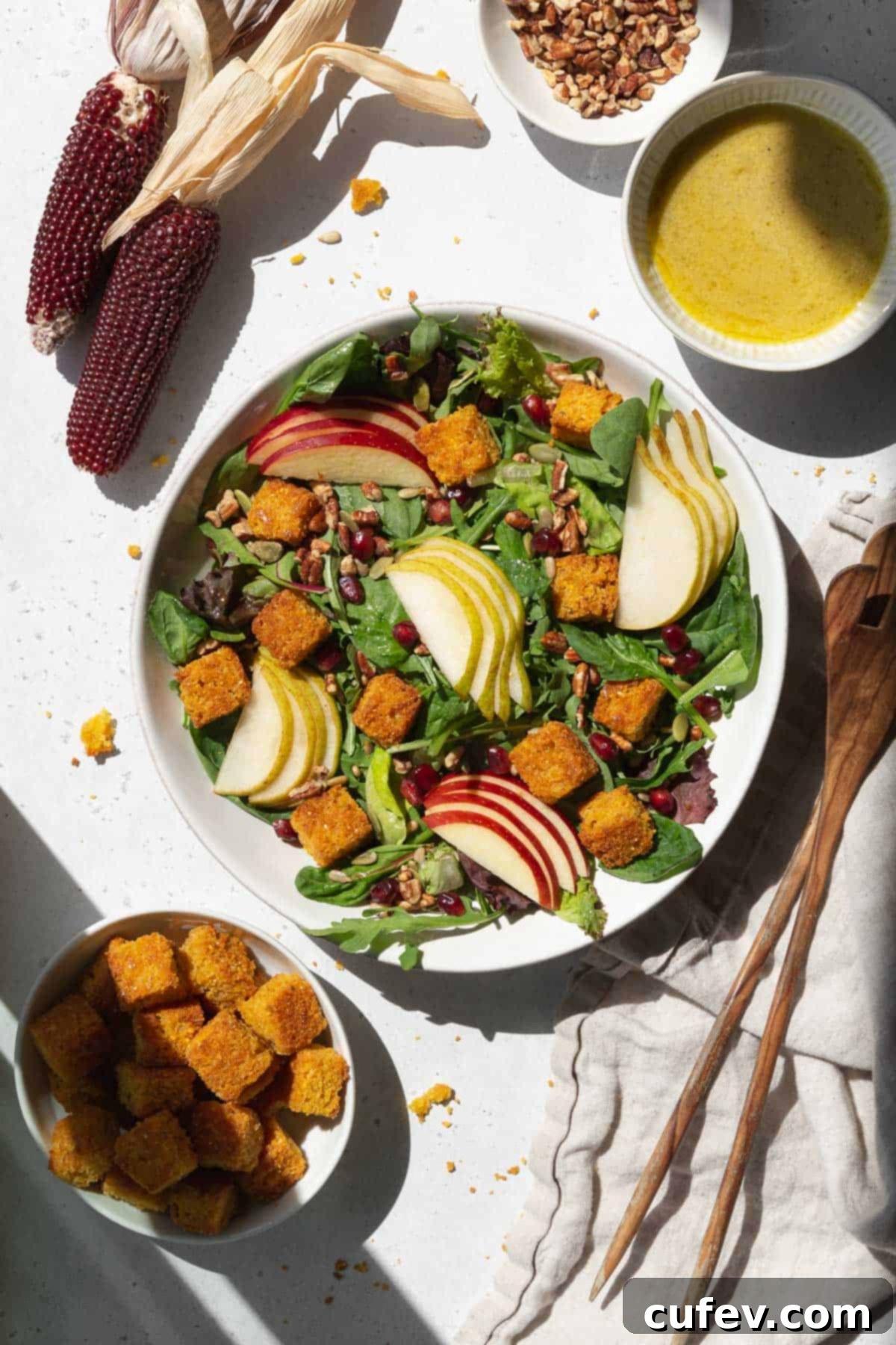 A vibrant spread for a salad, featuring a bowl of golden cornbread croutons, alongside sliced pears, apples, a dressing dispenser, and salad tongs.