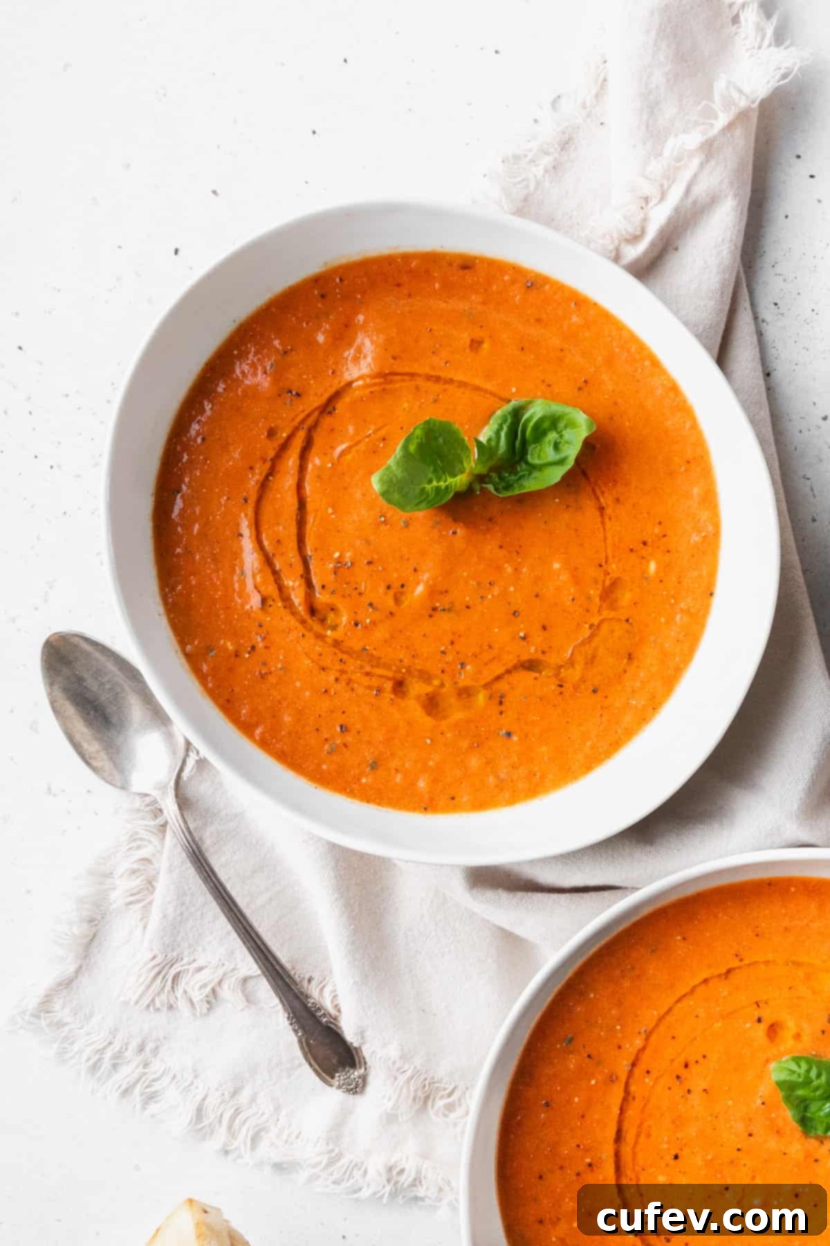 A white bowl of tomato basil soup with a sprig of basil on a linen and a silver spoon placed next to it.