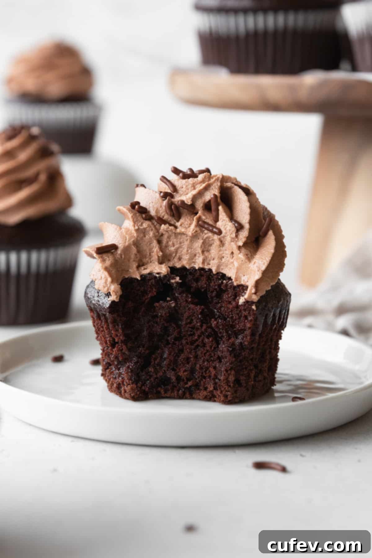 A close-up of a partially eaten chocolate cupcake with chocolate cream cheese frosting, revealing its moist crumb, resting on a pristine white plate.
