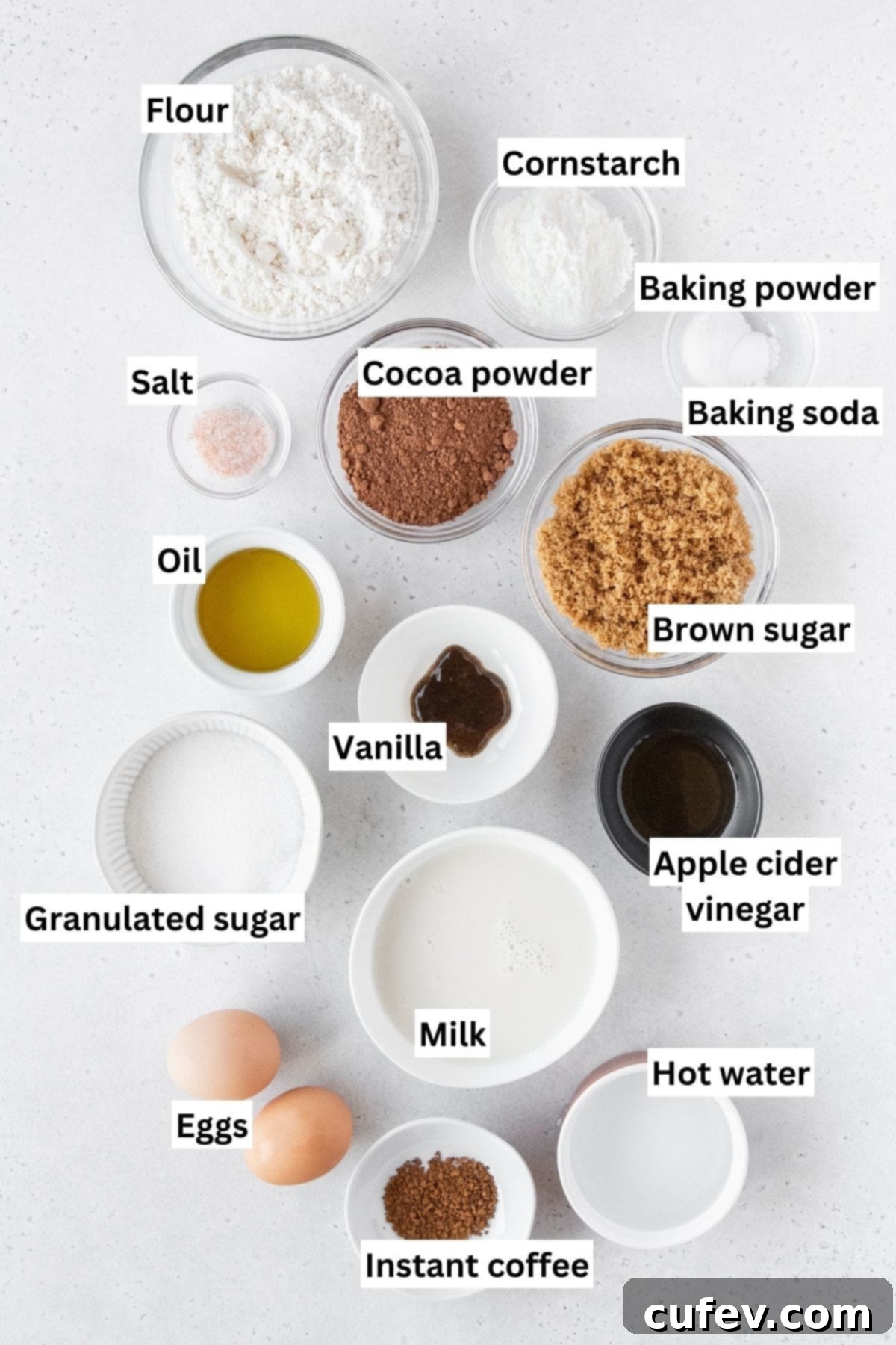 A collection of measured ingredients for chocolate cupcakes, including flours, sugars, cocoa, and liquids, neatly arranged in small bowls on a clean white surface, ready for baking.