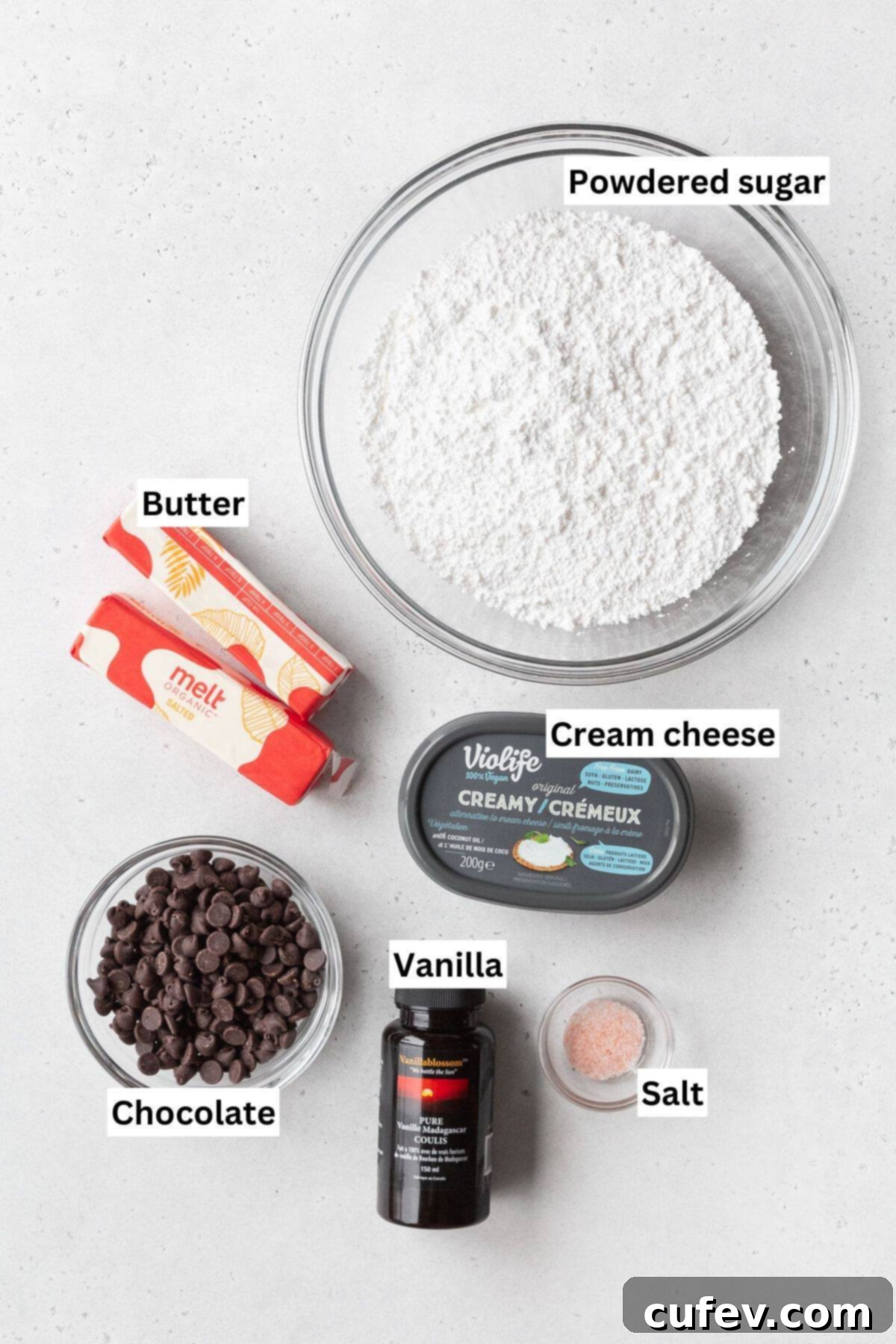 Various ingredients for a decadent chocolate cream cheese frosting, including softened butter, block cream cheese, powdered sugar, and melted semi-sweet chocolate, neatly prepared in bowls on a white background.
