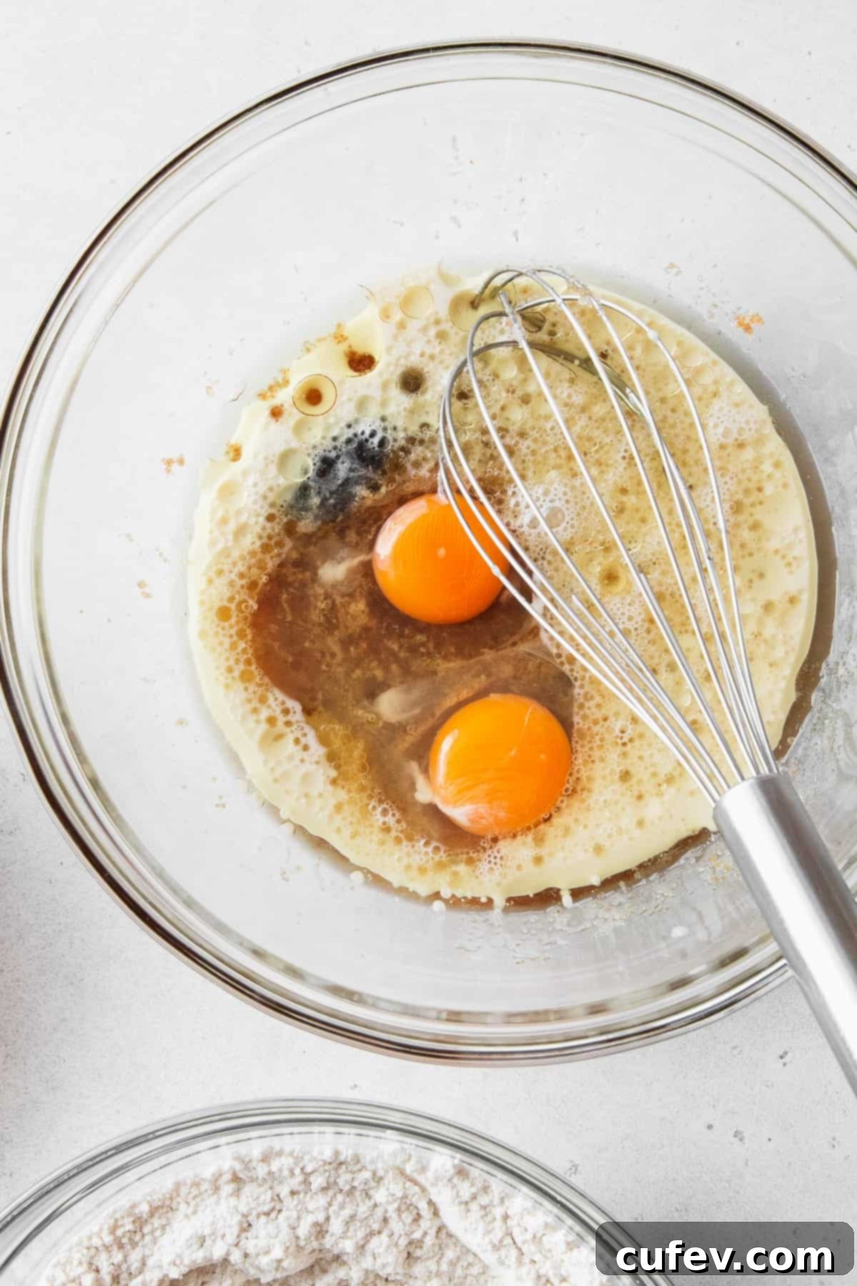 A hand whisking eggs, milk, apple cider vinegar, and vanilla extract into the sugar and oil mixture in a glass bowl, creating a smooth, light-colored wet batter.