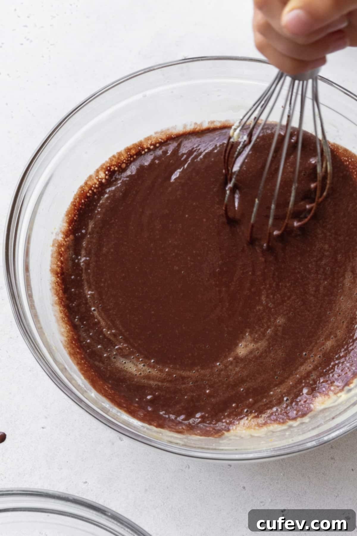 Rich, dark chocolate cupcake batter being expertly mixed with a whisk in a clear glass bowl, showing a smooth and consistent texture.