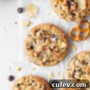 Kitchen sink cookie on a white surface surrounded by more cookies, chocolate chips, crushed potato chips, and pretzels.