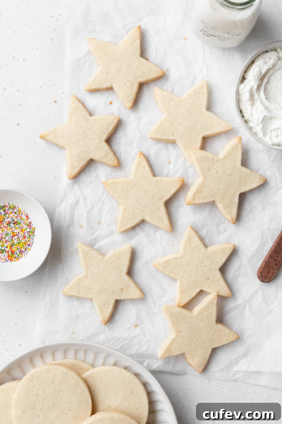 Star shaped gluten free sugar cookies on a white surface, with a small dish of sprinkles, a bowl of frosting, and a plate of circular sugar cookies on a plate surrounding them.