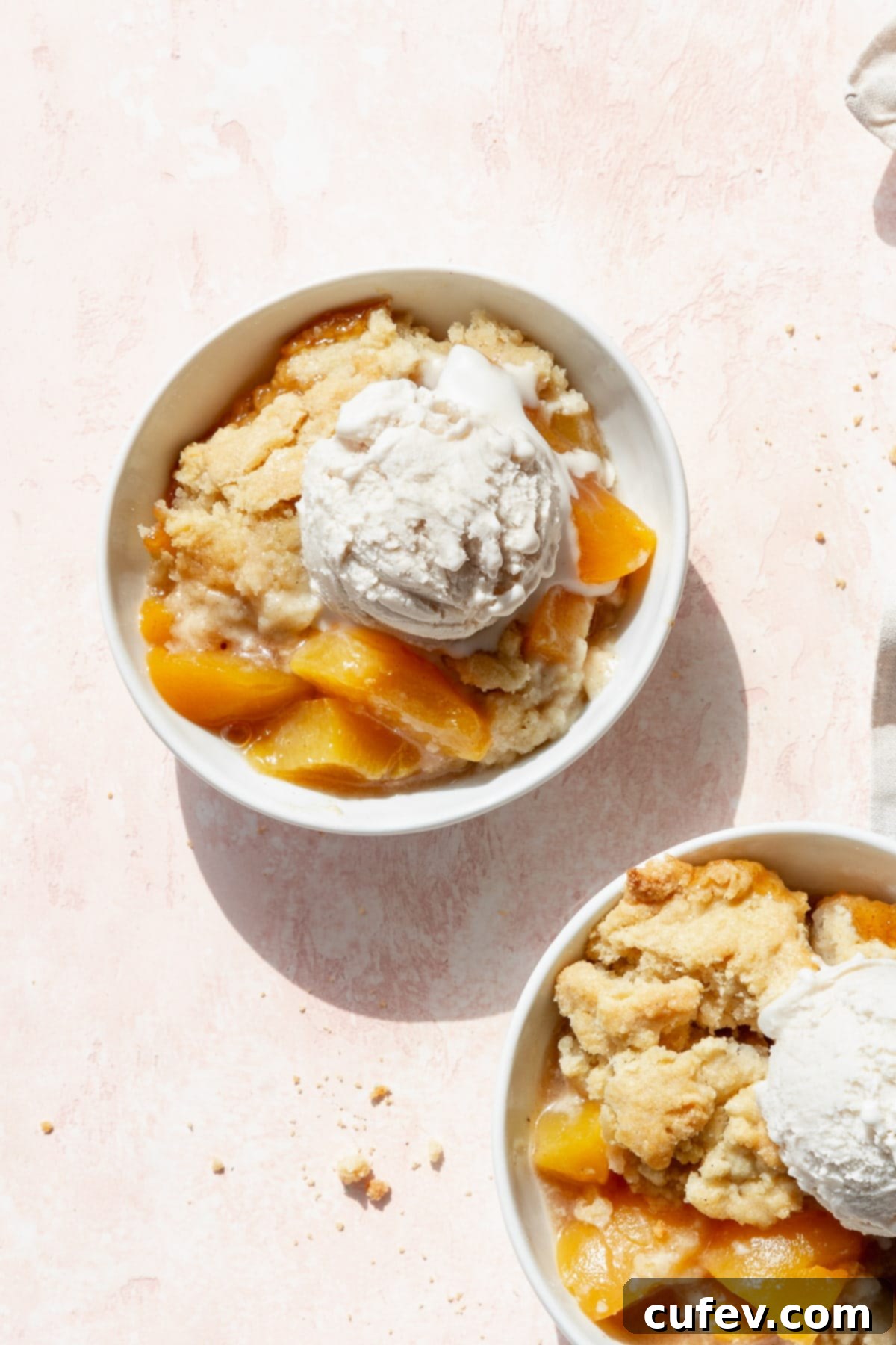 A bowl of peach cobbler with canned peaches and a scoop of vanilla ice cream on a blush colored surface.
