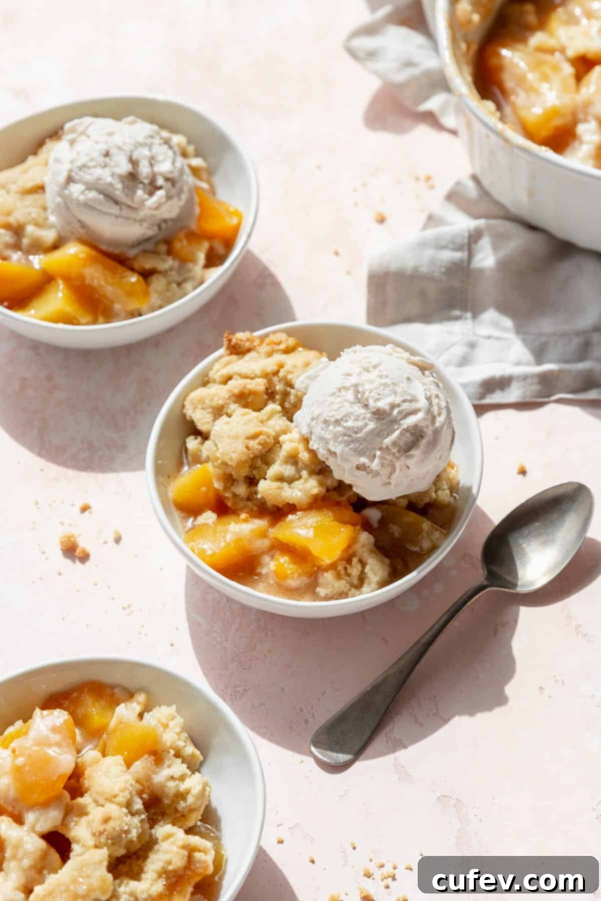 Peach cobbler with a scoop of ice cream in a dessert bowl with a spoon next to it, and more bowls scattered around.