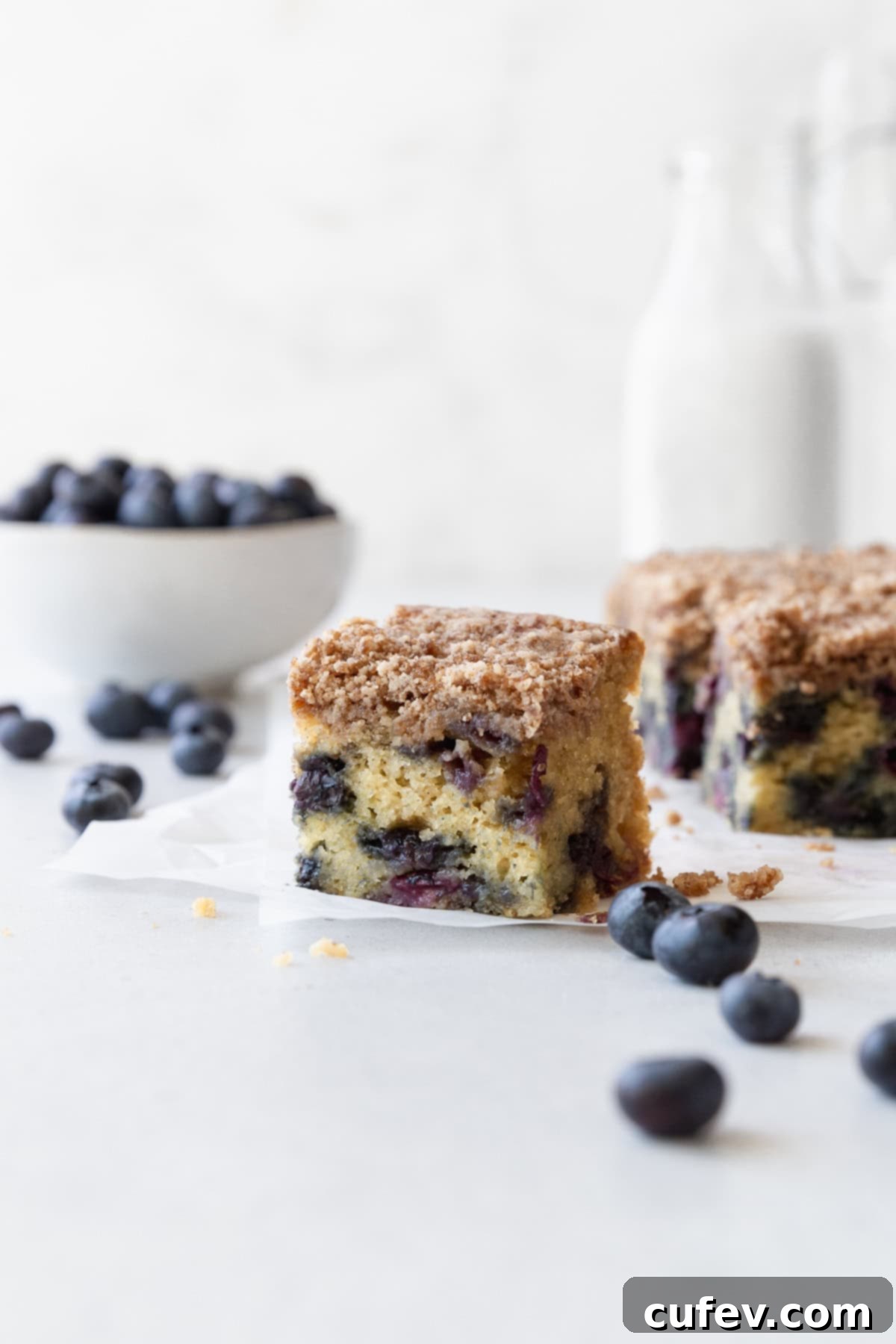 A square of blueberry coffee cake on a white surface with fresh blueberries scattered around.