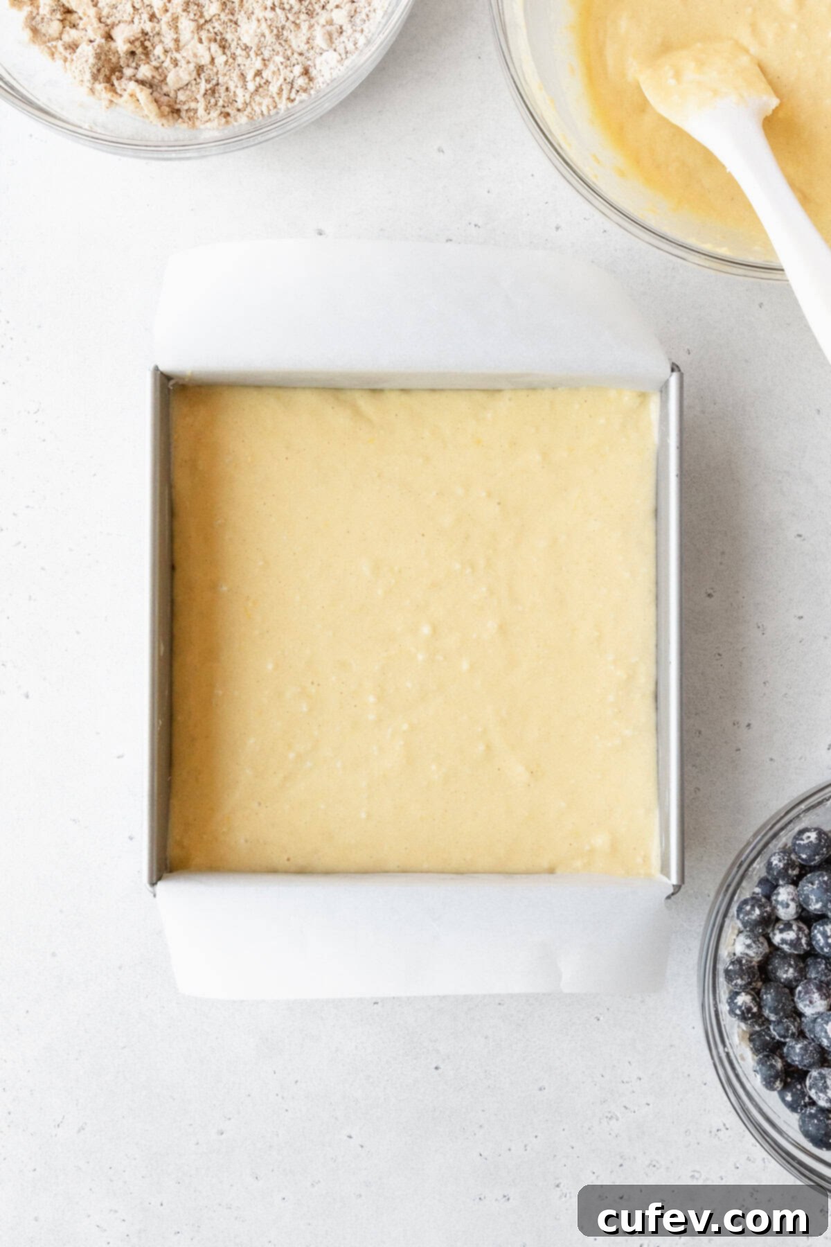 Cake batter in a square pan surrounded by more bowls of ingredients.
