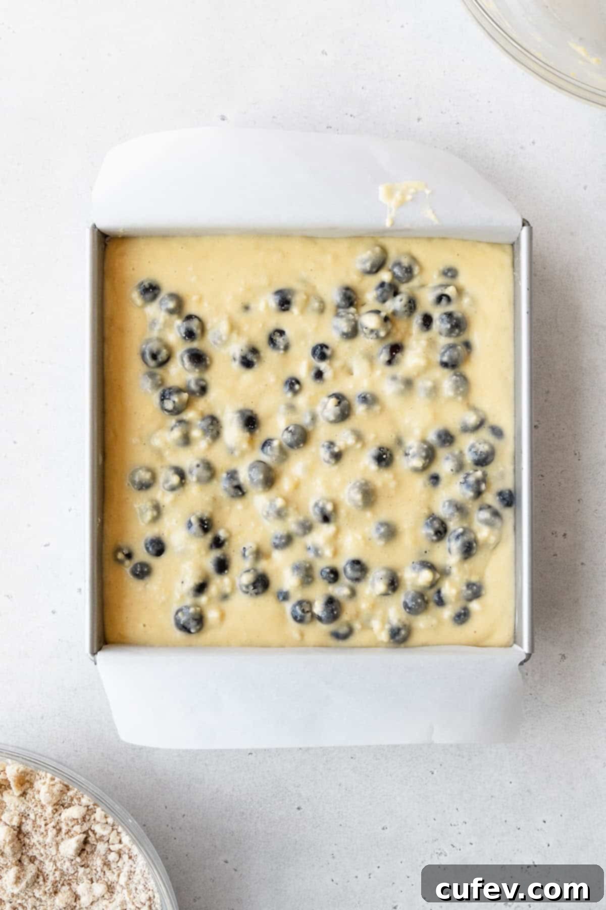 Blueberry cake batter in a square pan on a white surface.