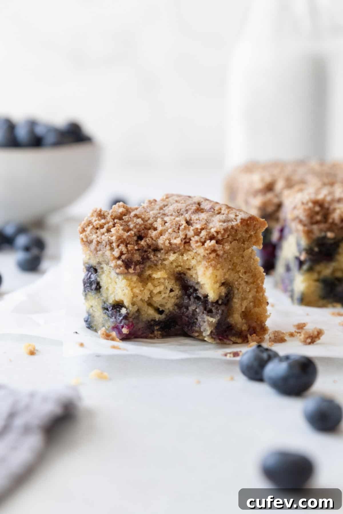 A blueberry coffee cake square with a bite taken out, with a bottle of milk and a bowl of blueberries in the background.