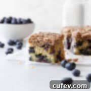 A slice of blueberry coffee cake on a white surface with blueberries scattered.