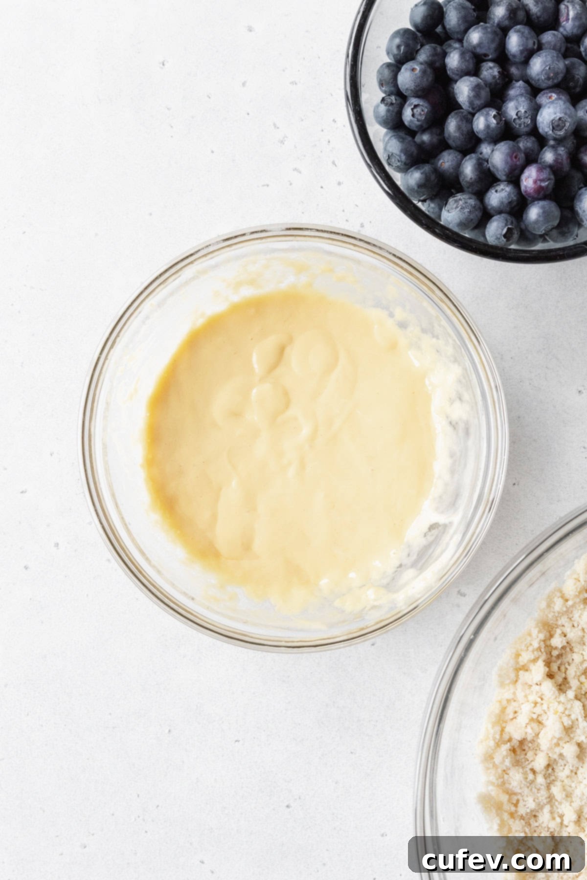 Wet ingredients mixed in a glass bowl with a bowl of blueberries diagonally above it.
