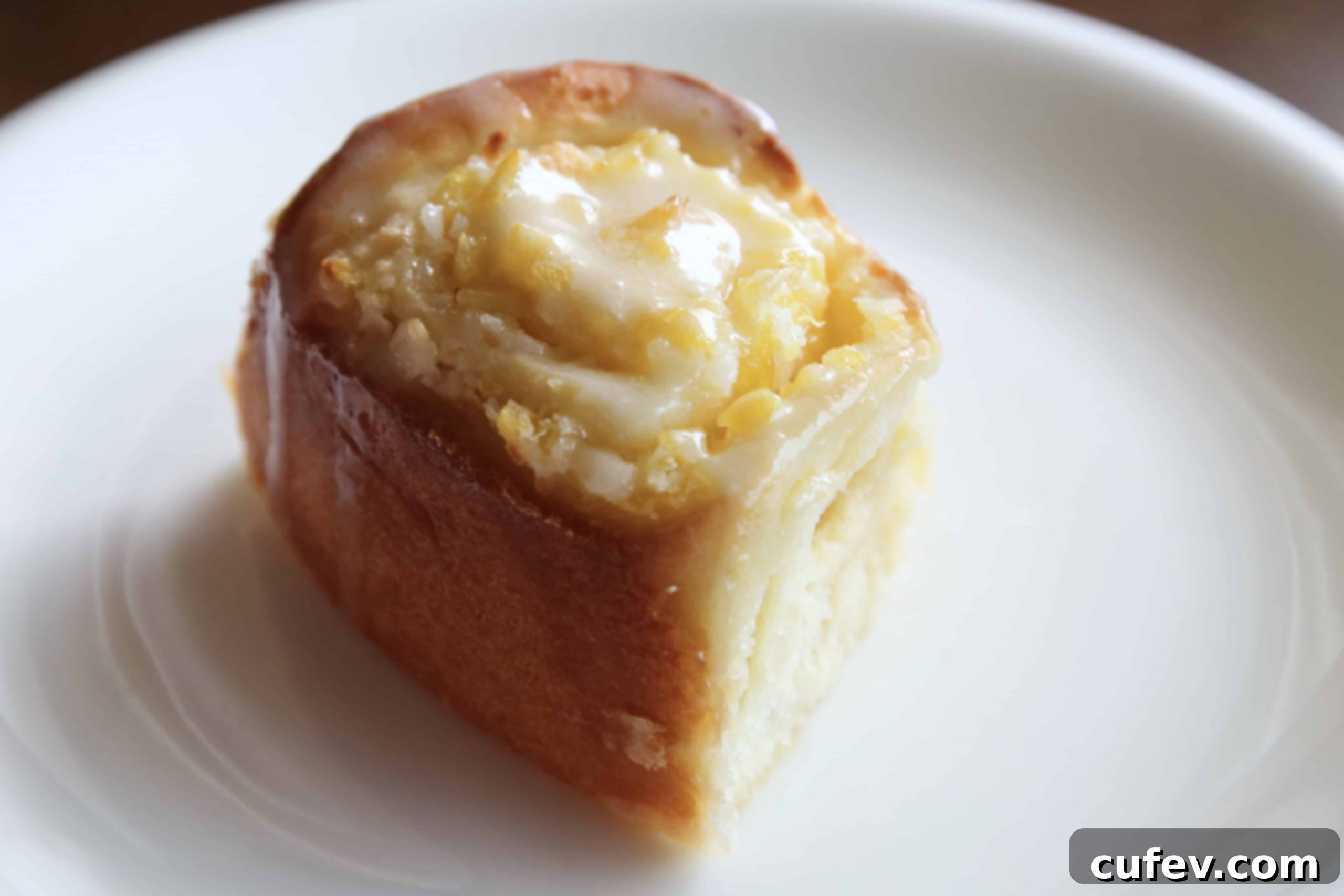 A single Piña Colada roll on a white plate, showcasing its golden-brown crust, visible filling, and creamy white frosting.